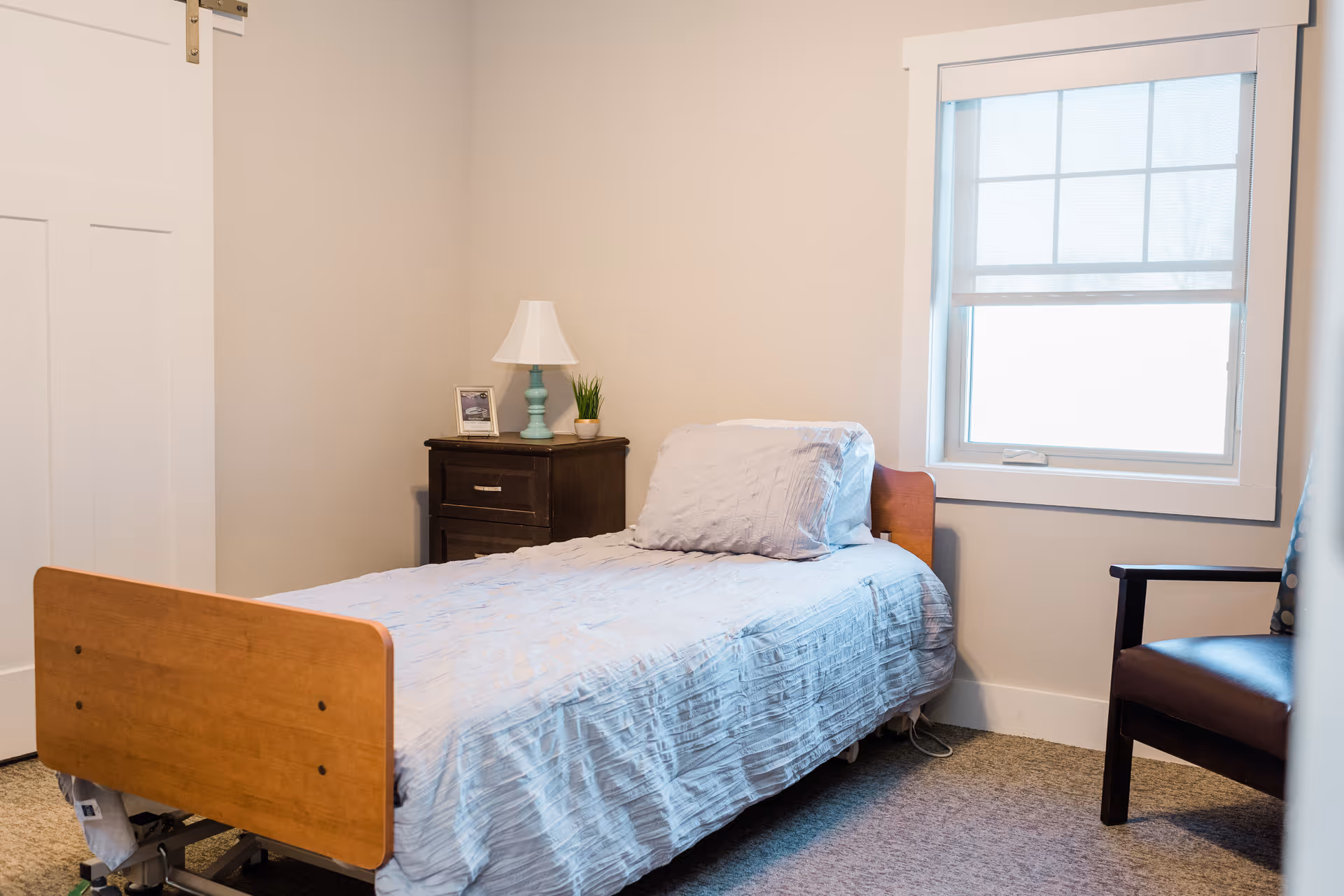 Small, tidy bedroom with a single wooden hospital-style bed, a nightstand with a lamp and plant, a window with a shade, and an armchair.