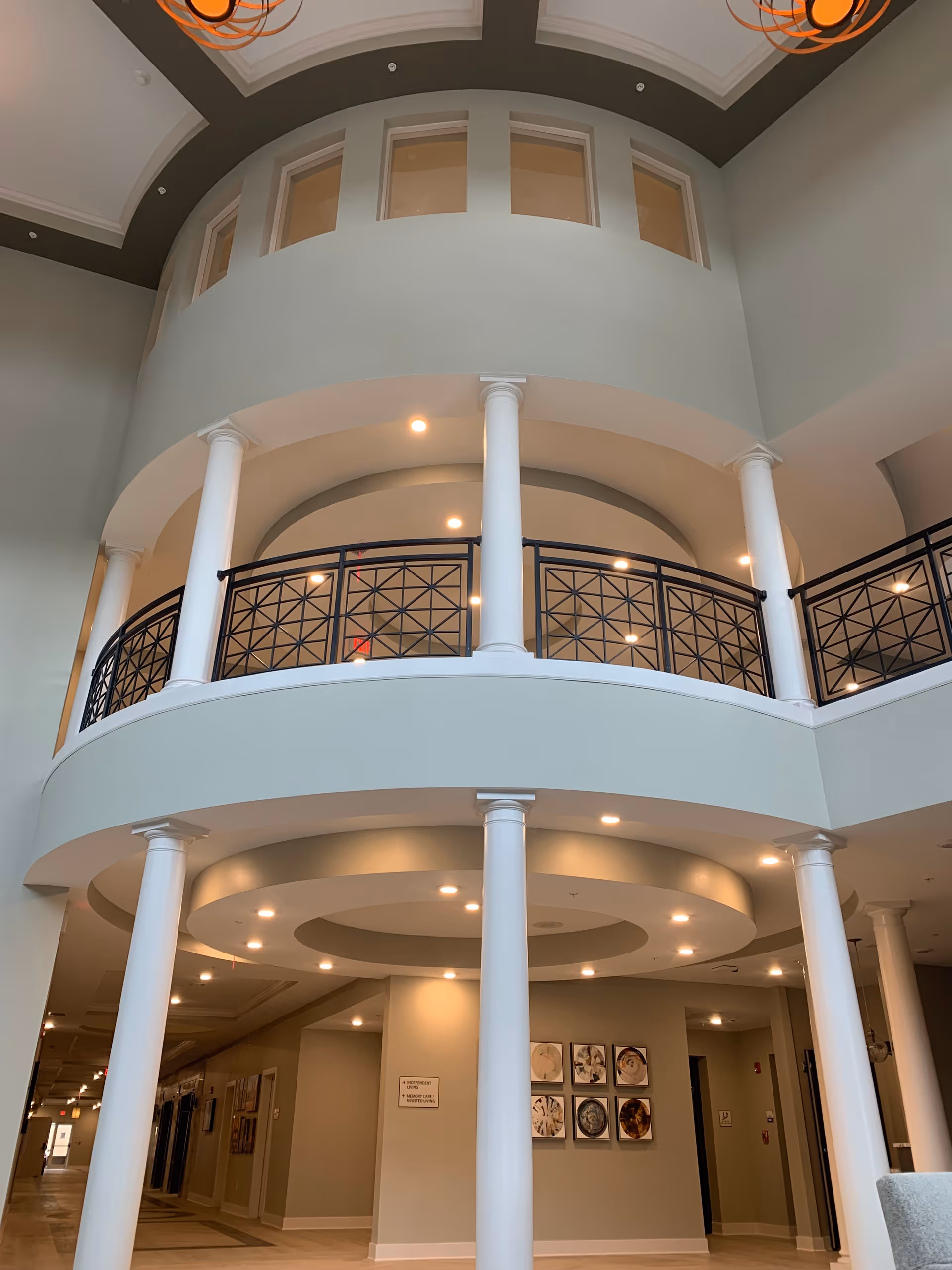 Interior view of a senior living facility with a two-story circular balcony supported by white columns. The ceiling has recessed lighting and decorative molding. There are framed pictures on the wall and a hallway leading to other rooms.