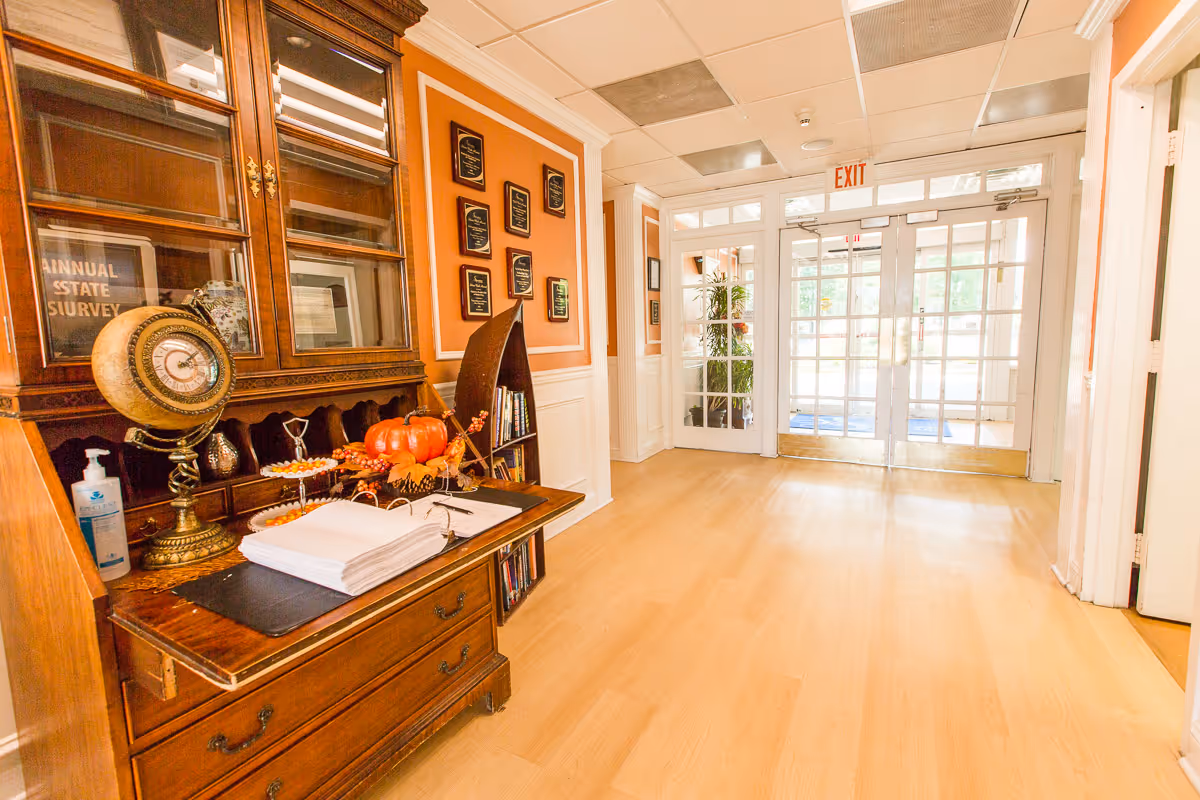 Bright hallway with light wood flooring and white trim leading to glass double doors with an exit sign above. On the left side, there is a wooden cabinet with decorative items including a globe, a pumpkin, and a hand sanitizer bottle. Several plaques are mounted on the orange wall above a small bookshelf.
