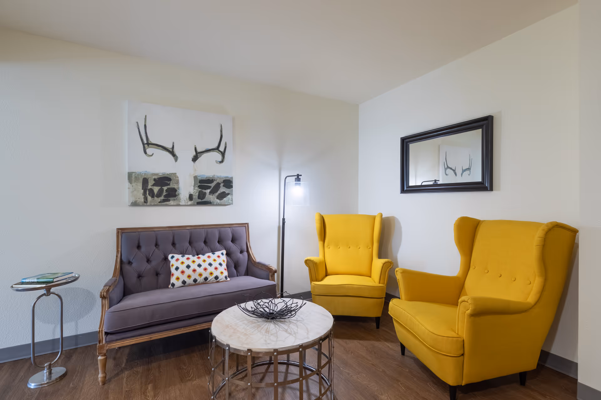 A cozy sitting area with two yellow wingback chairs, a tufted purple loveseat with a colorful patterned pillow, a round marble-top coffee table with a decorative wire bowl, a floor lamp, a small side table with books, and a wall mirror reflecting part of the room and artwork featuring antlers.