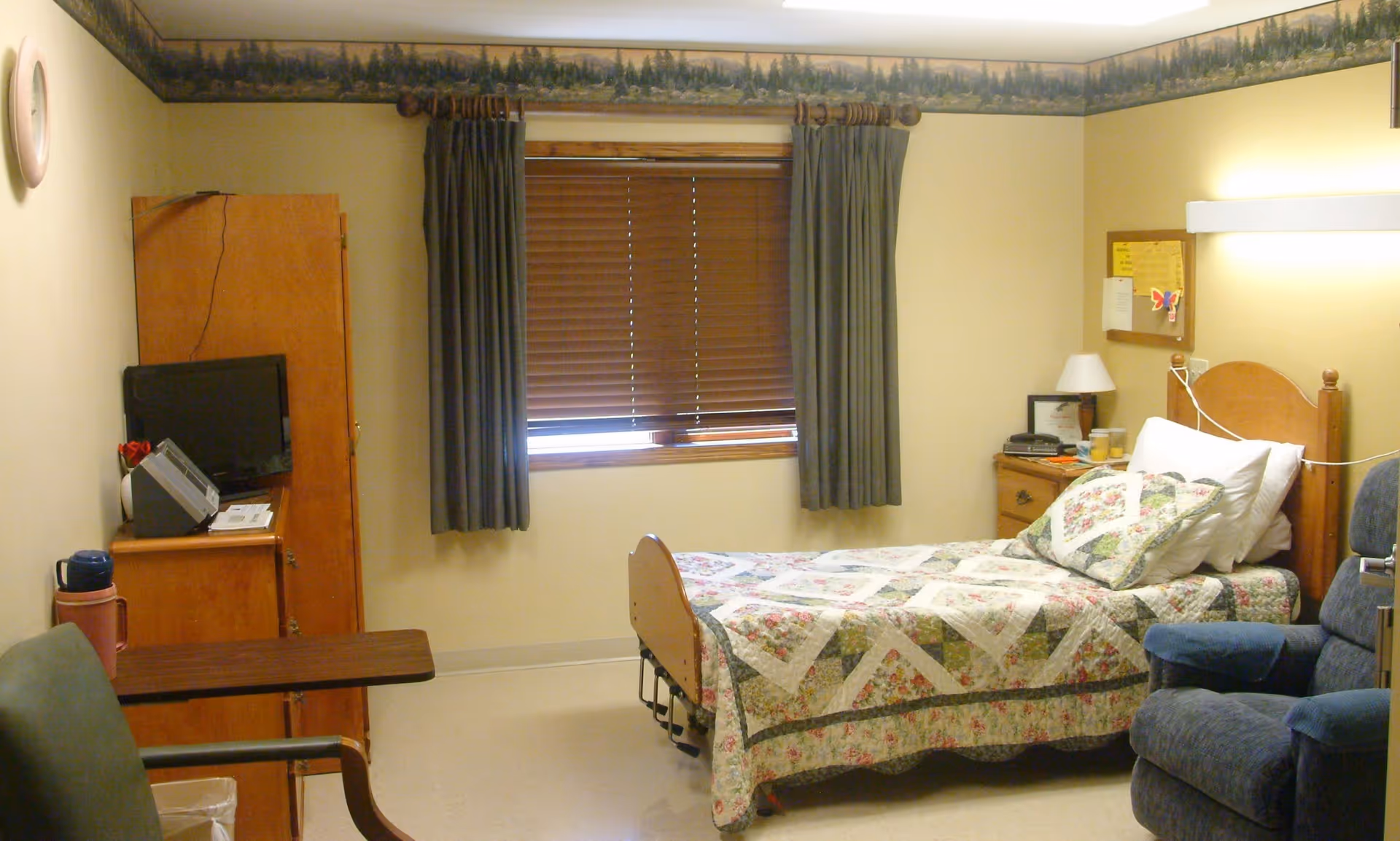 A small, cozy bedroom in a senior living facility with a single bed covered in a floral quilt. The room has beige walls with a nature-themed border near the ceiling, a window with closed wooden blinds and dark curtains, a wooden nightstand with a lamp and some items, a blue upholstered armchair, a small wooden table, and a cabinet with a TV on top.