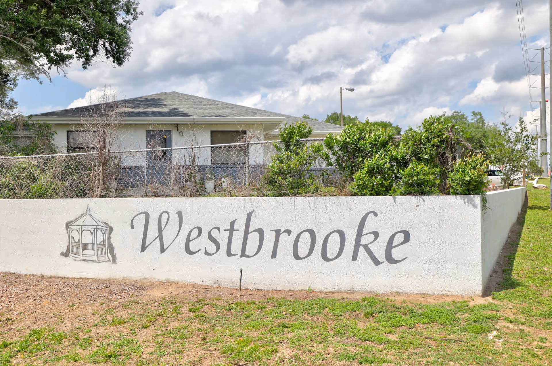 Exterior view of a single-story building behind a white concrete wall with the word 'Westbrooke' painted on it alongside an illustration of a gazebo. There is a chain-link fence and some bushes behind the wall, with a partly cloudy sky overhead.