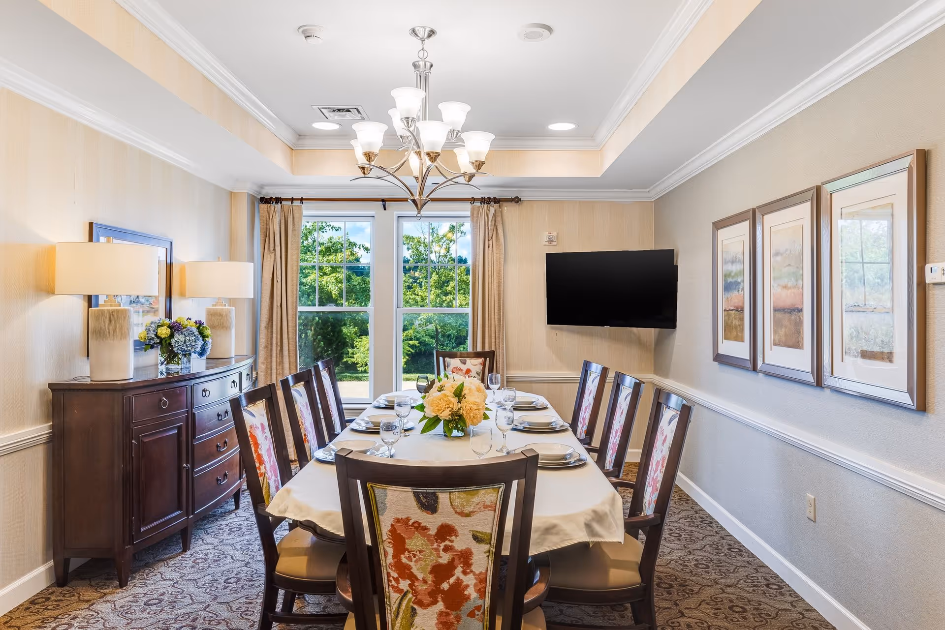 A well-lit dining room with a long table set for eight people. The table is covered with a white tablecloth and has plates, glasses, and a floral centerpiece. There are eight wooden chairs with floral upholstery around the table. The room features a chandelier, two large windows with beige curtains, a dark wooden sideboard with two lamps and a flower arrangement, a wall-mounted flat-screen TV, and three framed artworks on the wall.
