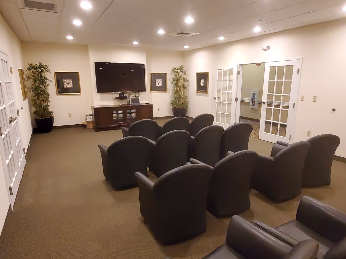A small theater-style room with two rows of black armchairs facing a wall-mounted flat screen TV. The room has beige walls, carpeted floor, two potted plants, framed artwork on the walls, and double glass doors leading to another area.