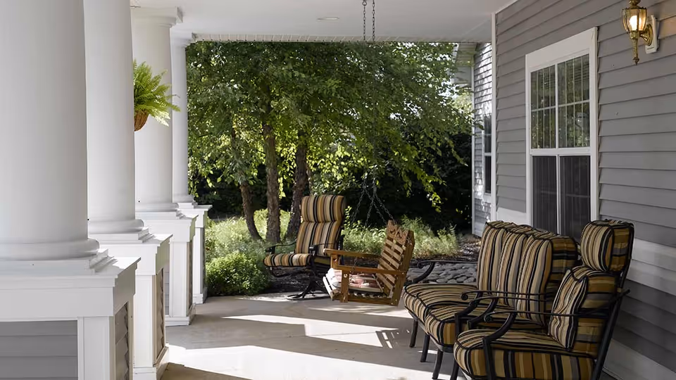 A covered outdoor porch area with white columns, cushioned striped chairs, a wooden swing, and greenery in the background.
