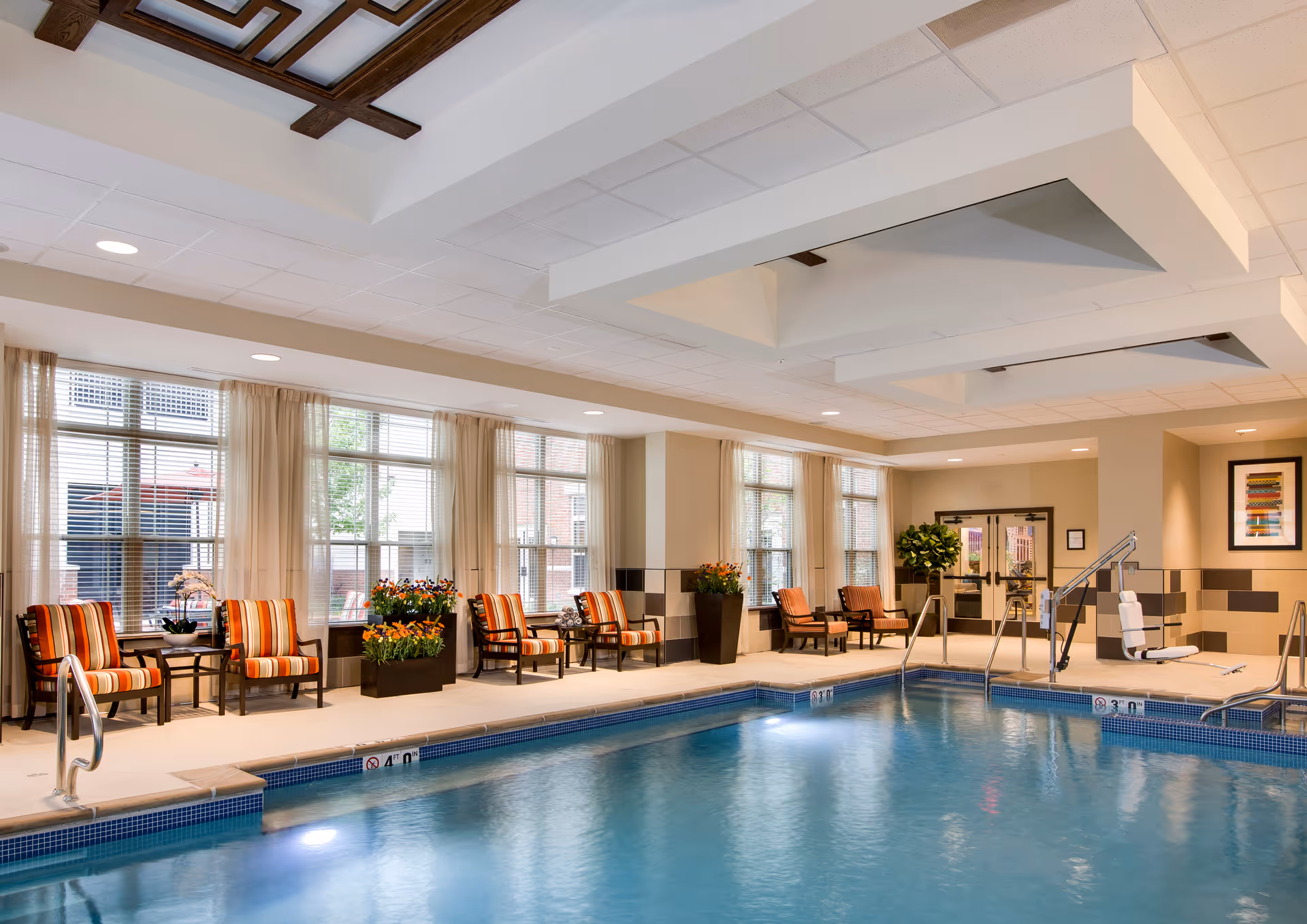Indoor swimming pool area with clear blue water, surrounded by a tiled deck. Several striped cushioned chairs and small tables with flower arrangements are placed along the windows. Large windows with beige curtains allow natural light to fill the space. The ceiling has recessed lighting and decorative wooden panels. A pool lift is installed for accessibility near the pool entrance.
