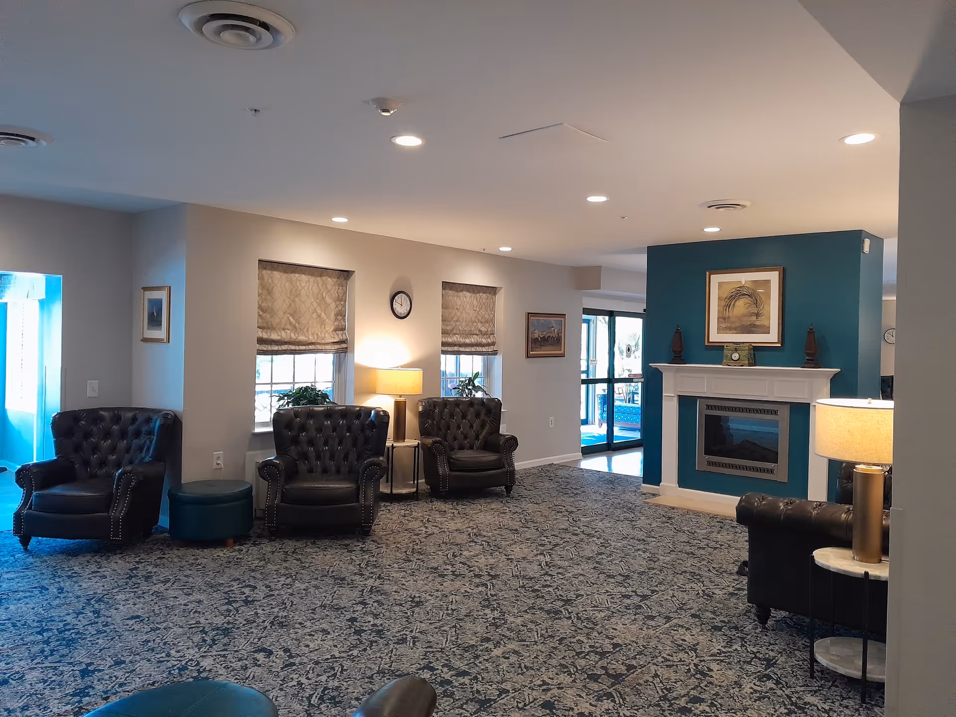 A cozy living room area with dark leather armchairs arranged around a fireplace with a teal accent wall. The room has patterned carpet, two windows with beige roman shades, a clock on the wall, framed artwork, and table lamps providing warm lighting. A glass door leads outside in the background.