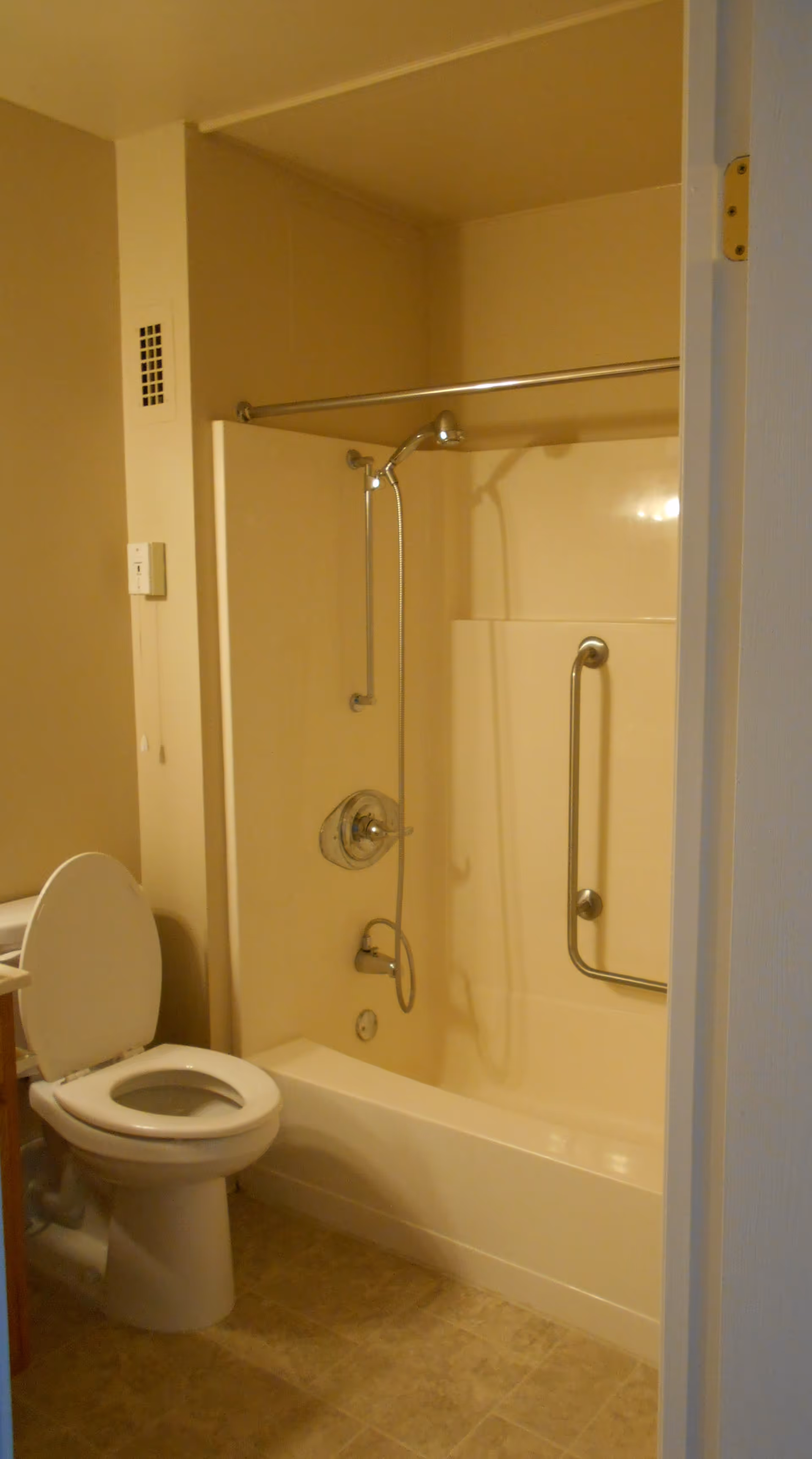 A bathroom with a white toilet with the lid open, a bathtub with a showerhead, a grab bar on the wall, and a shower curtain rod above the tub. The floor has beige tiles and the walls are painted a light color.