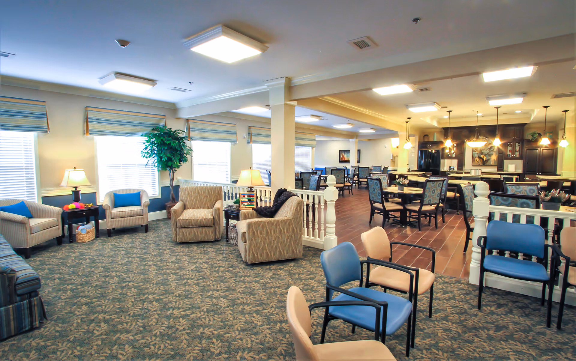 A spacious senior living facility common area with a seating lounge featuring armchairs and a sofa on a patterned carpet. Adjacent to the lounge is a dining area with multiple tables and chairs, pendant lights hanging from the ceiling, and a kitchen area with dark cabinetry and appliances in the background.
