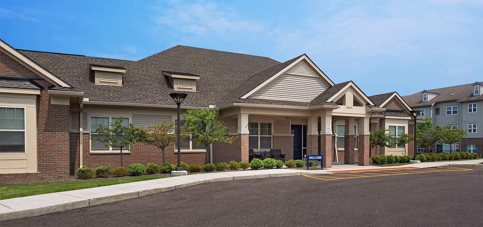 Exterior view of a single-story brick and siding building with a covered entrance, small trees, shrubs, and a paved driveway under a clear blue sky.