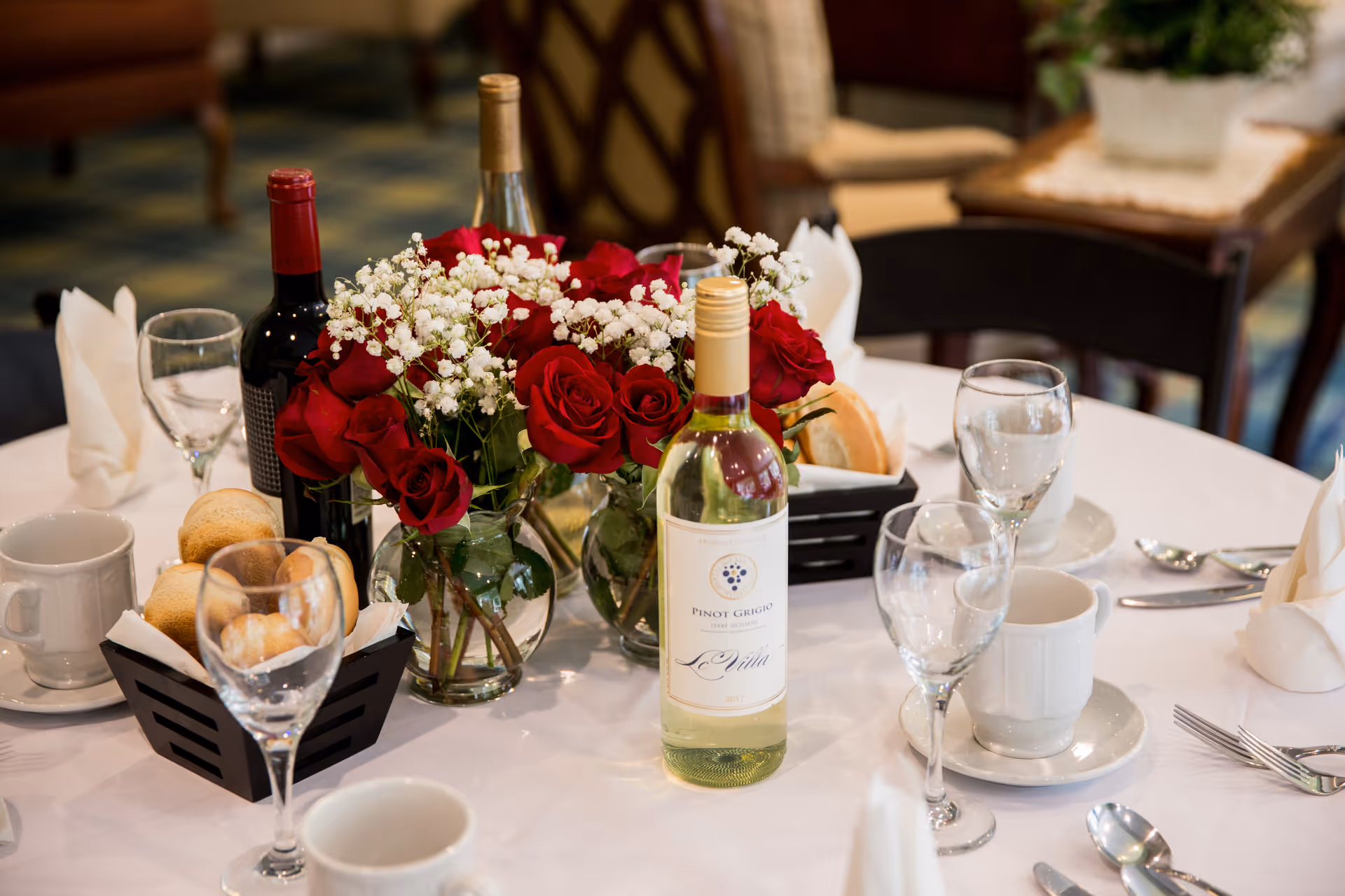 A round dining table set with white tablecloth, wine bottles, glasses, coffee cups, bread rolls in baskets, and a centerpiece of red roses and white baby's breath flowers in glass vases.