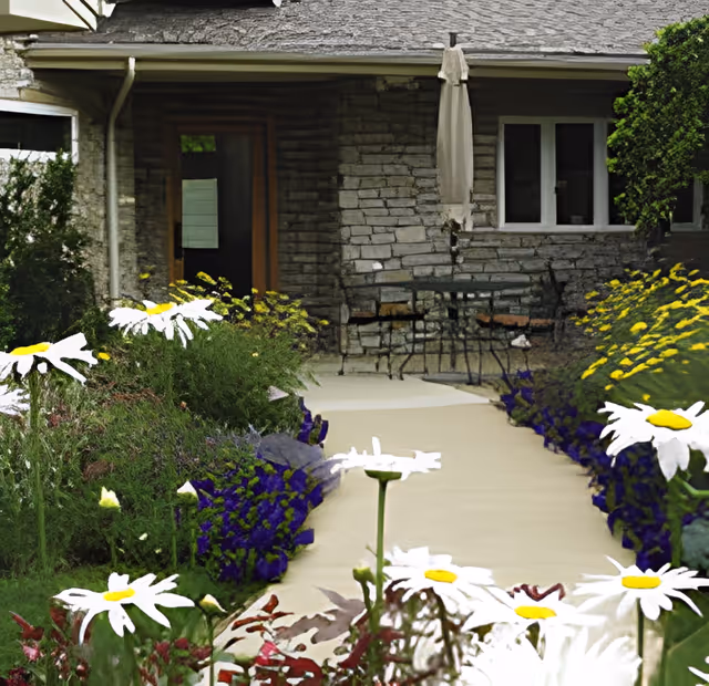 A stone building with a pathway leading to a patio area with a table and chairs. The pathway is bordered by colorful flowers including white daisies and purple and yellow blooms. There is a closed umbrella near the table and windows on the building wall.