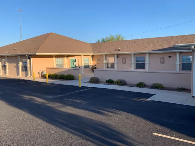 Exterior view of a single-story senior living facility with a pink-beige facade, brown roof, and a parking lot in front.