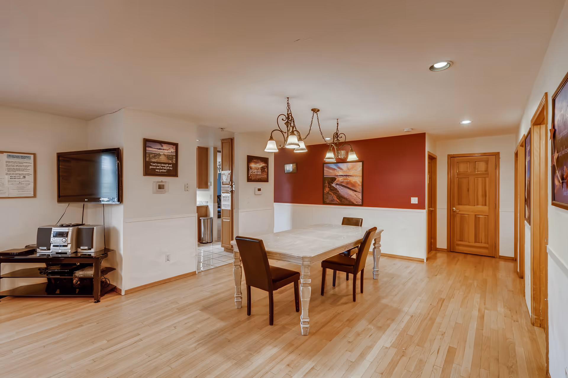 Bright dining room with a long wooden table, four chairs, a chandelier, wall art, and a TV/stereo on a stand.