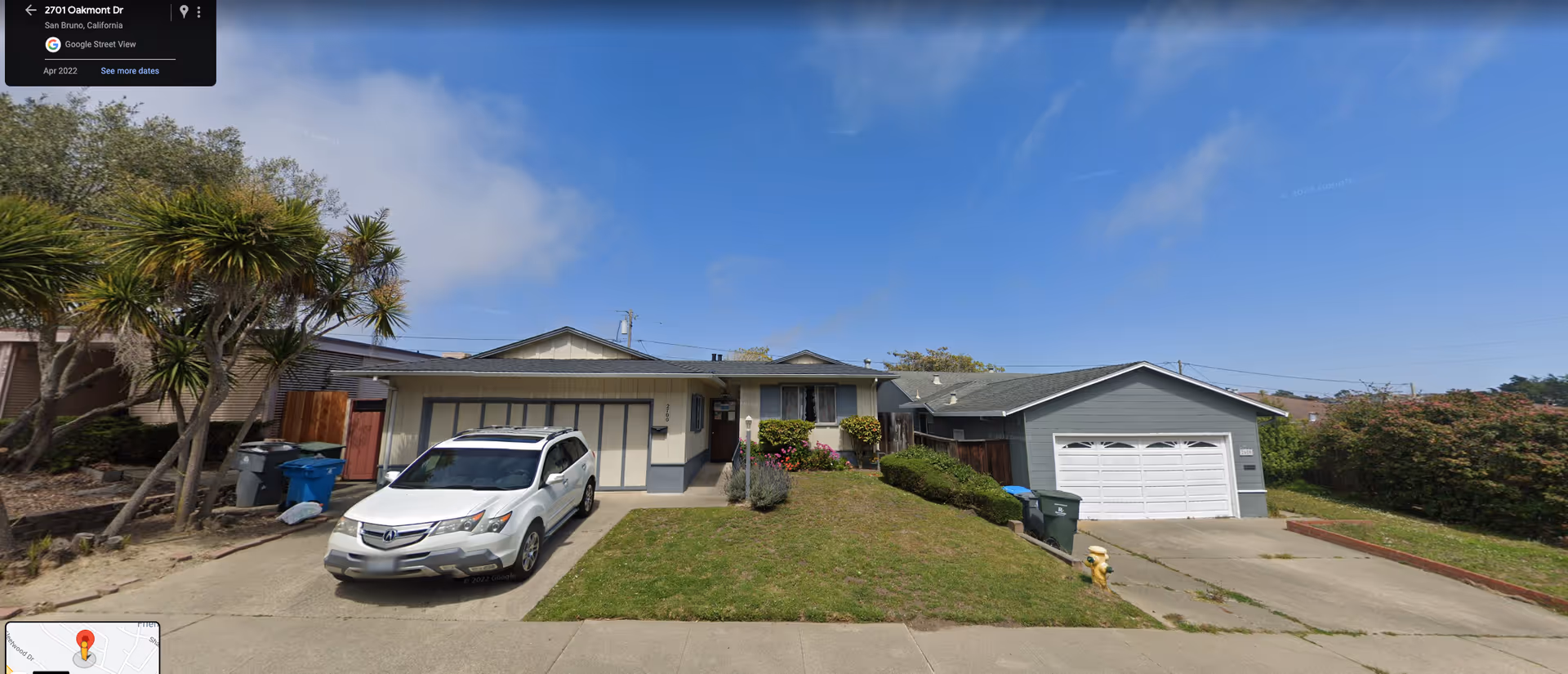 Front exterior view of a single-story residential house with a driveway and a white SUV parked in front. The house has a garage with closed doors, a small lawn with bushes and flowers, and a clear blue sky above.