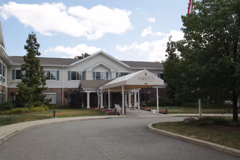 Front exterior view of a two-story senior living facility building with a covered entrance labeled 'Emeritus'. The building is surrounded by trees and landscaping under a partly cloudy sky.