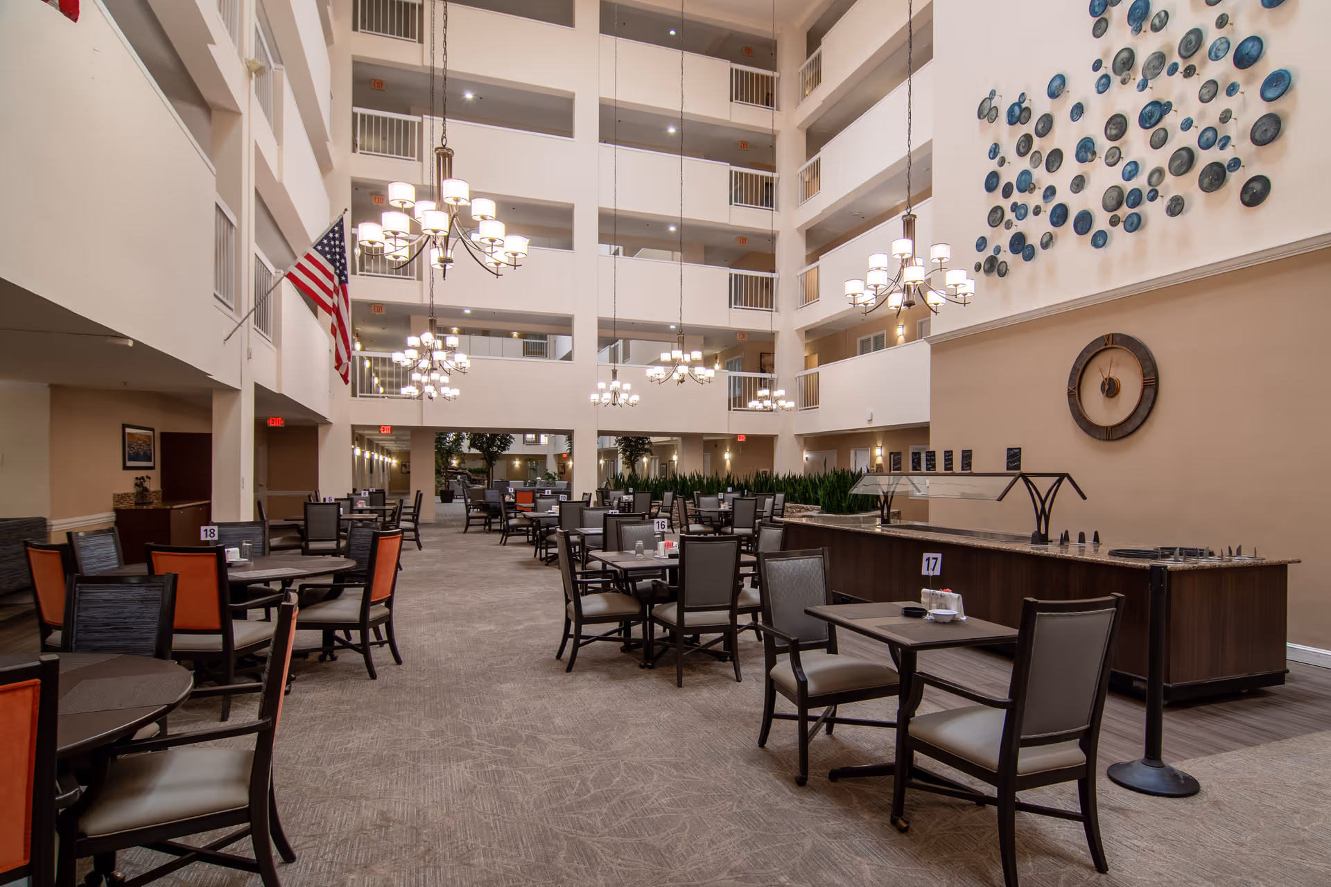 Spacious dining area inside Dunwoody Pines Retirement Community with multiple tables and chairs arranged neatly. The room features high ceilings with several hanging chandeliers, an American flag on the left wall, and a decorative wall clock and blue circular wall art on the right. There is a buffet counter along the right side and multiple floors with balconies overlooking the dining area.