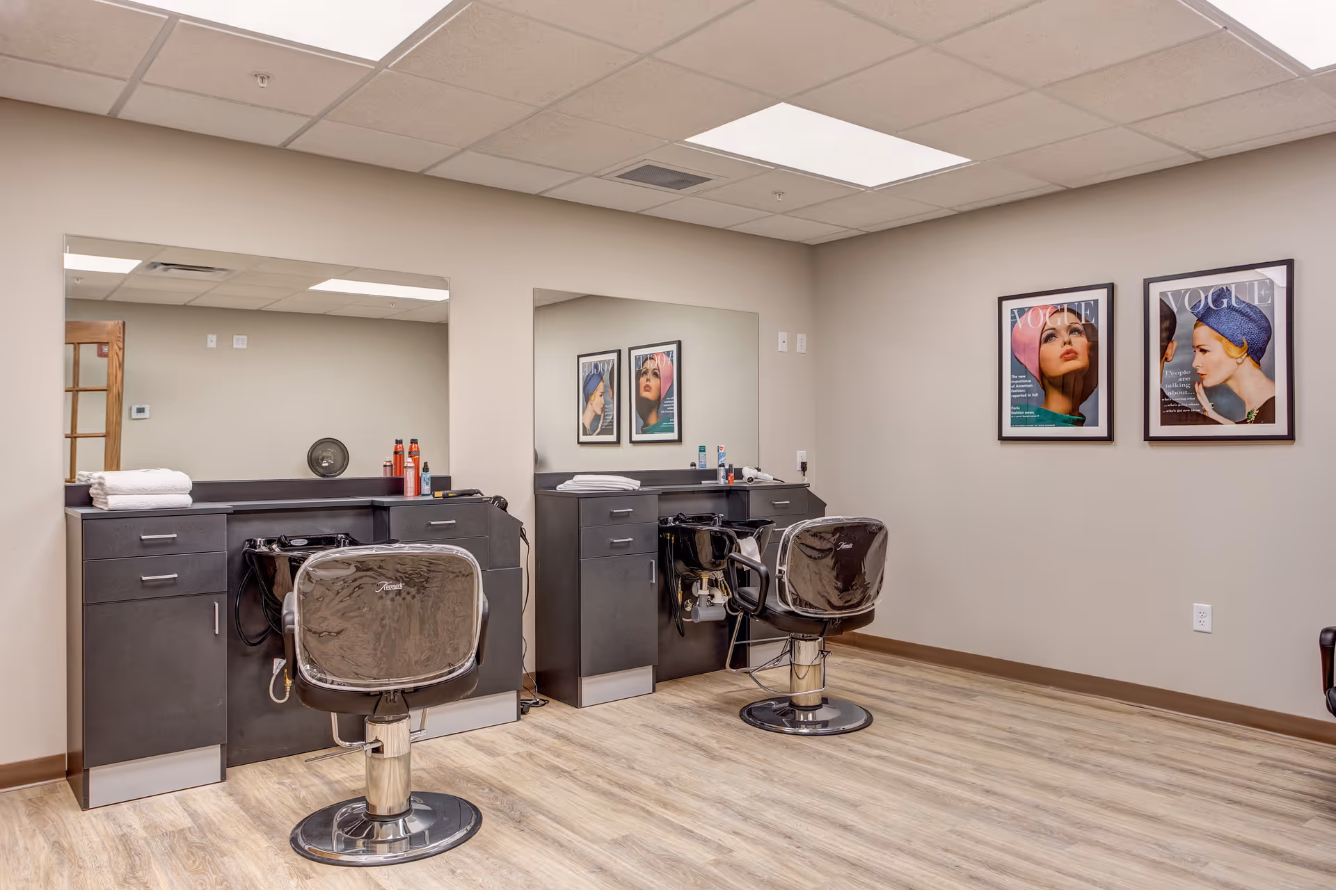 Interior view of a hair salon area in a retirement community with two black salon chairs facing large mirrors mounted on beige walls. The salon stations have black cabinets with hair care products and towels on the countertops. Two framed Vogue magazine covers are hung on the wall to the right. The floor is light wood, and the ceiling has recessed lighting panels.