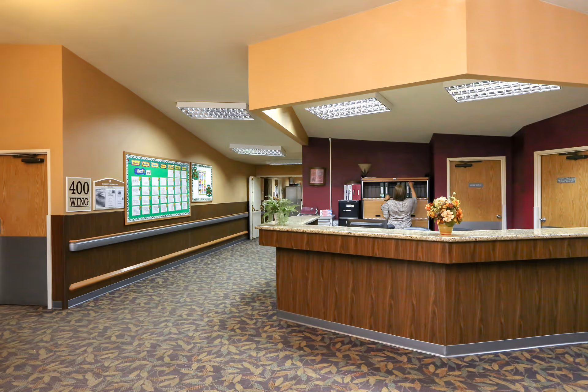Interior view of a senior living facility reception area with a wooden front desk, a person organizing files behind the desk, a flower arrangement on the counter, bulletin boards on the wall, and doors leading to other rooms. The floor is carpeted with a leaf pattern and the walls are painted in warm tones.
