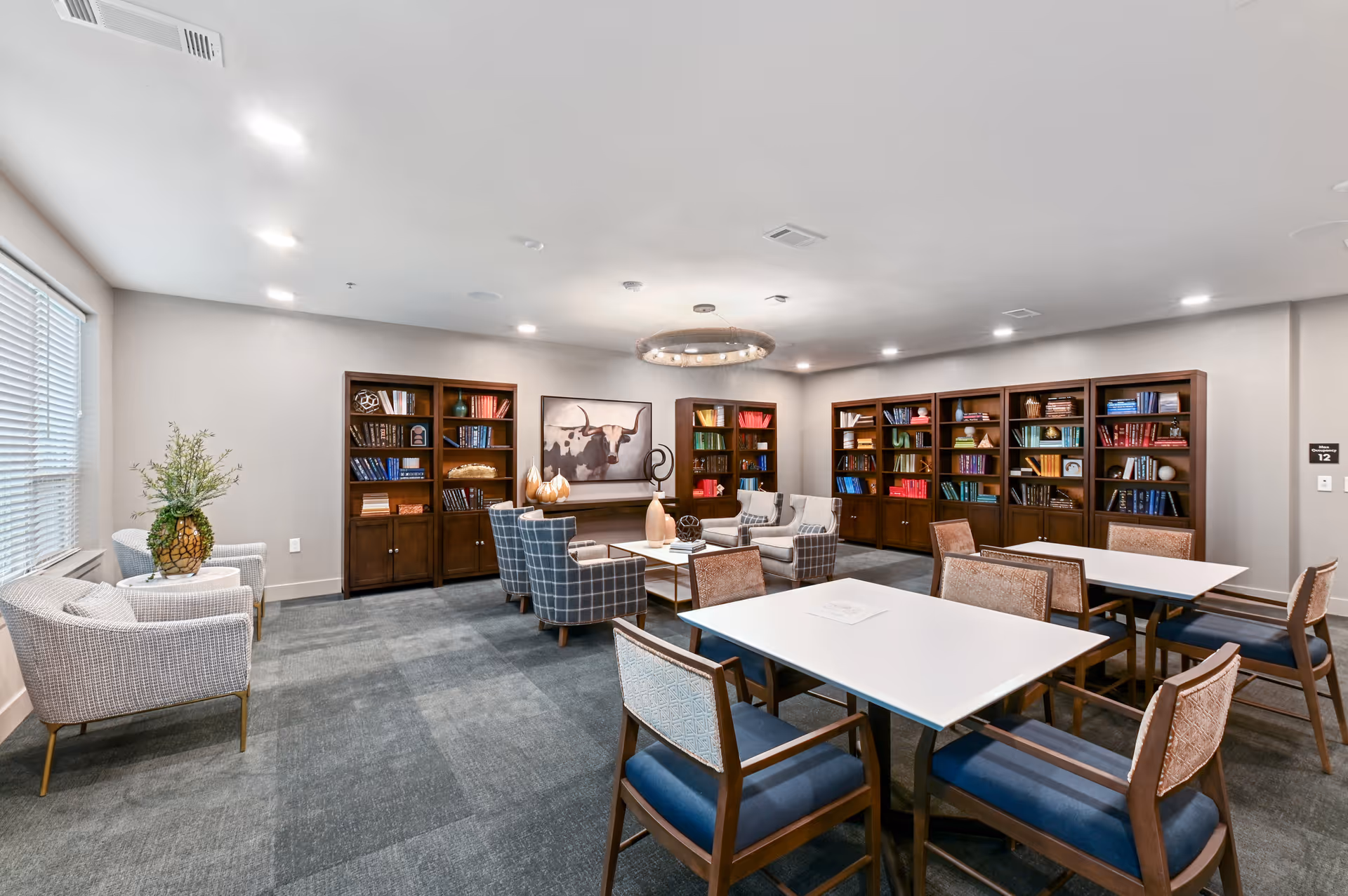 A well-lit common area in a senior living facility featuring multiple bookshelves filled with books and decorative items along the walls. There are several seating arrangements including armchairs and tables with chairs, a large window with blinds on the left, and a modern circular chandelier hanging from the ceiling.