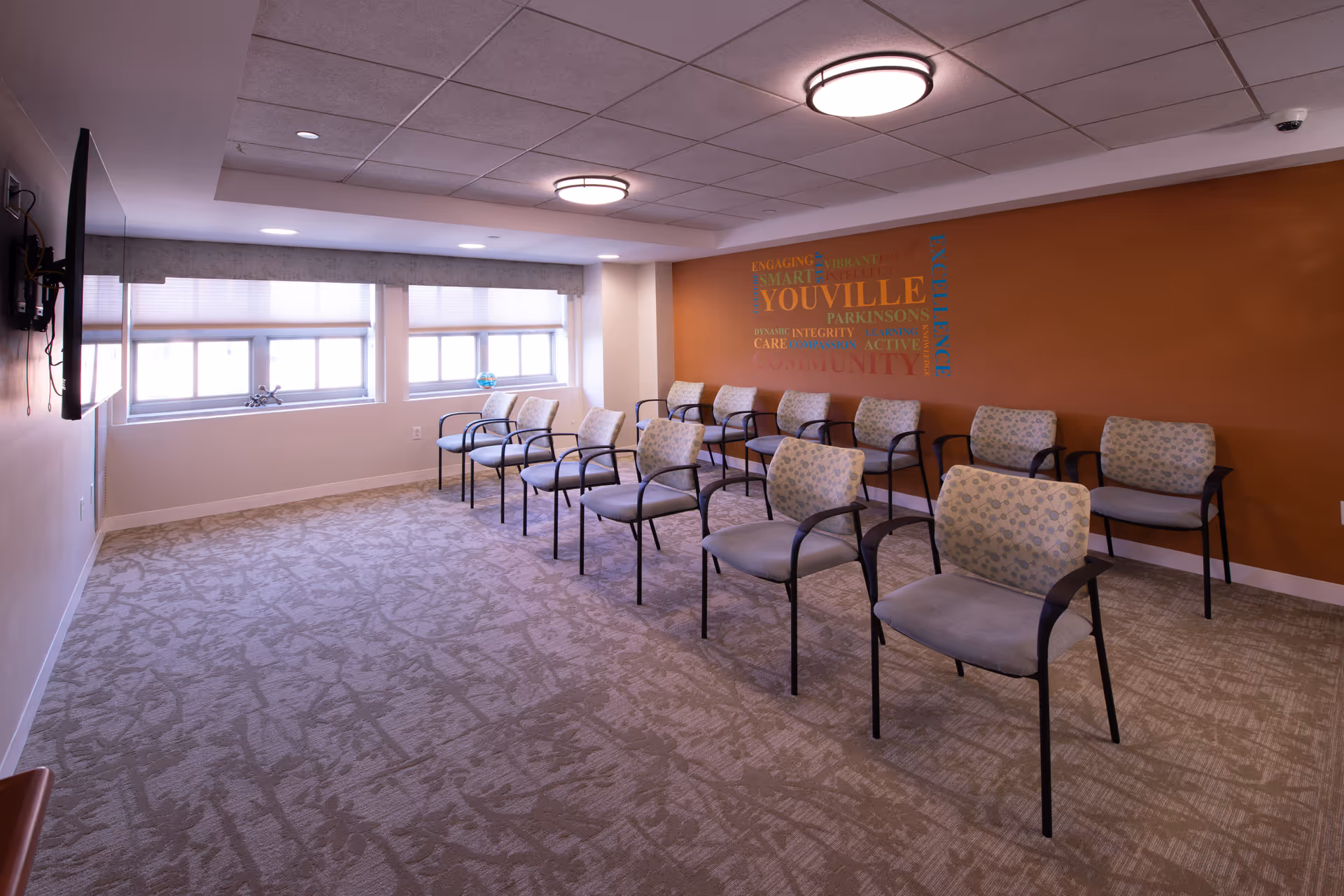 A small meeting/activity room with rows of chairs facing a wall-mounted TV and 'Youville' word art on an orange accent wall.