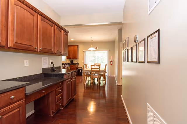 Interior view of a residential assisted living facility showing a kitchen area with wooden cabinets and black countertops on the left, leading to a dining area with a wooden table and chairs near a window with natural light.