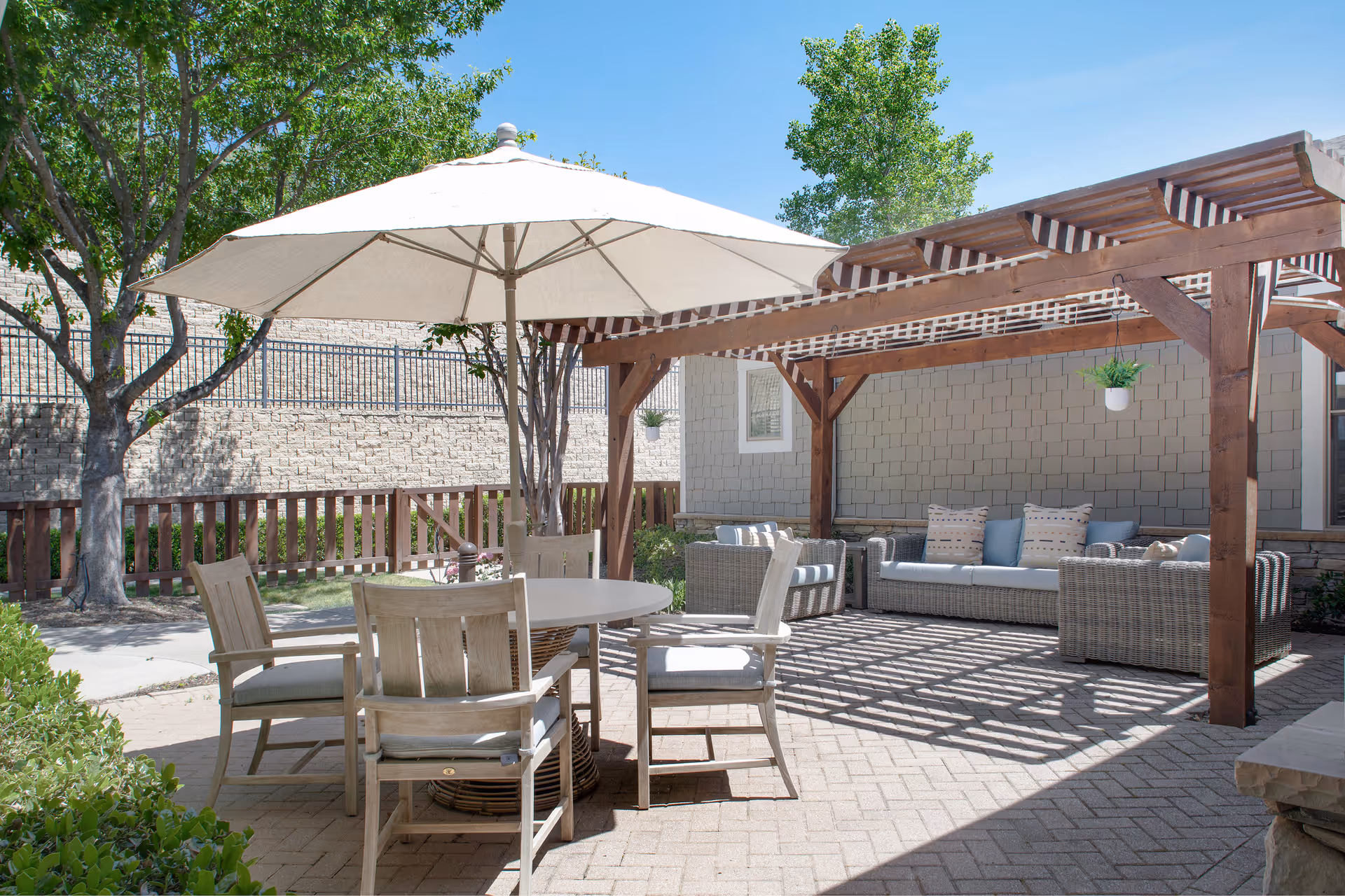 Outdoor patio area with a round table and four cushioned wooden chairs under a large white umbrella. There is a wooden pergola with hanging plants shading a wicker sofa and armchairs with cushions. Trees and a wooden fence surround the paved patio area under a clear blue sky.