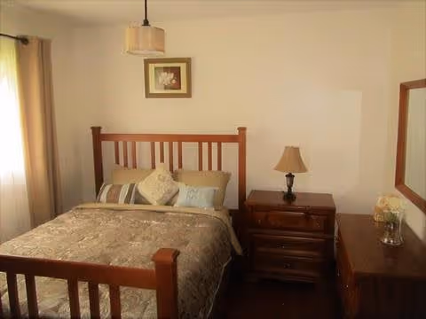 Well-lit bedroom with a wooden bed, nightstand and lamp, dresser with flowers, and framed art on the wall.