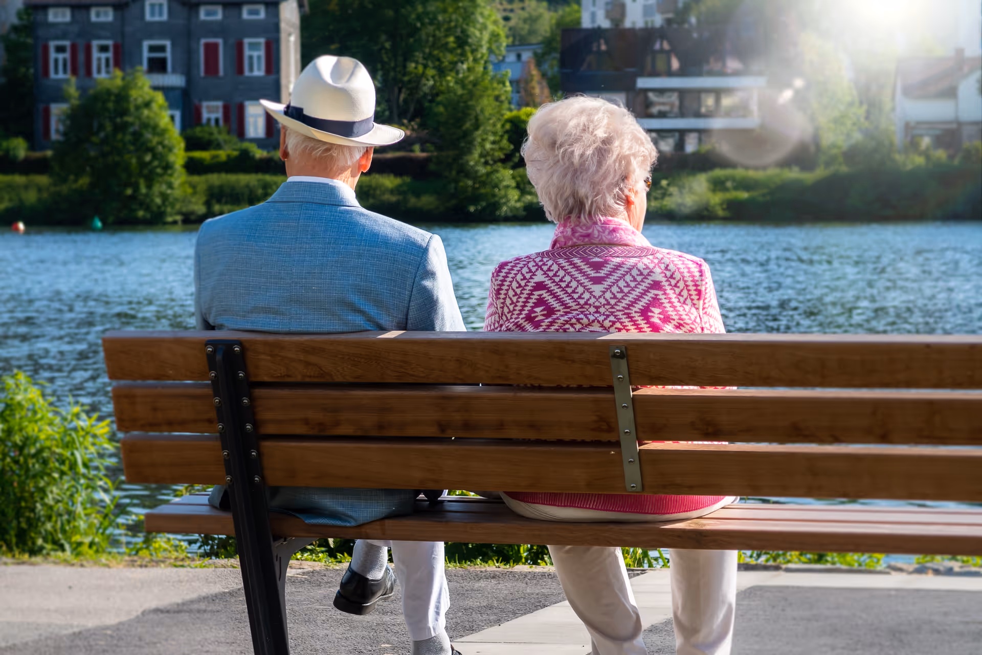 An elderly man wearing a white hat and blue jacket and an elderly woman wearing a pink patterned sweater sitting on a wooden bench by a lake with trees and houses in the background.