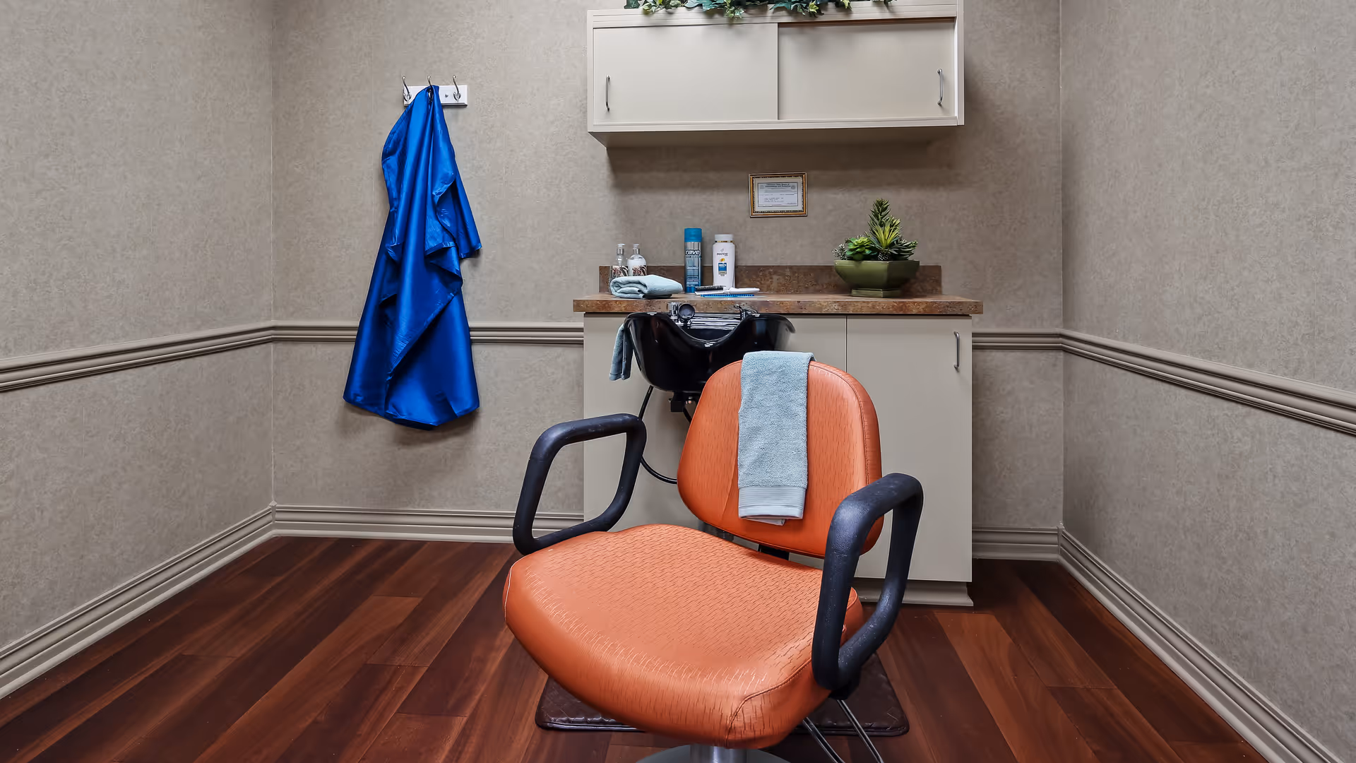 A small hair salon room with an orange salon chair in front of a black hair washing sink. A light blue towel is draped over the chair back. On the wall, a blue salon cape hangs on a hook. Behind the sink, there is a countertop with hair care products and a small green plant. Above the counter, there is a white cabinet mounted on the wall.