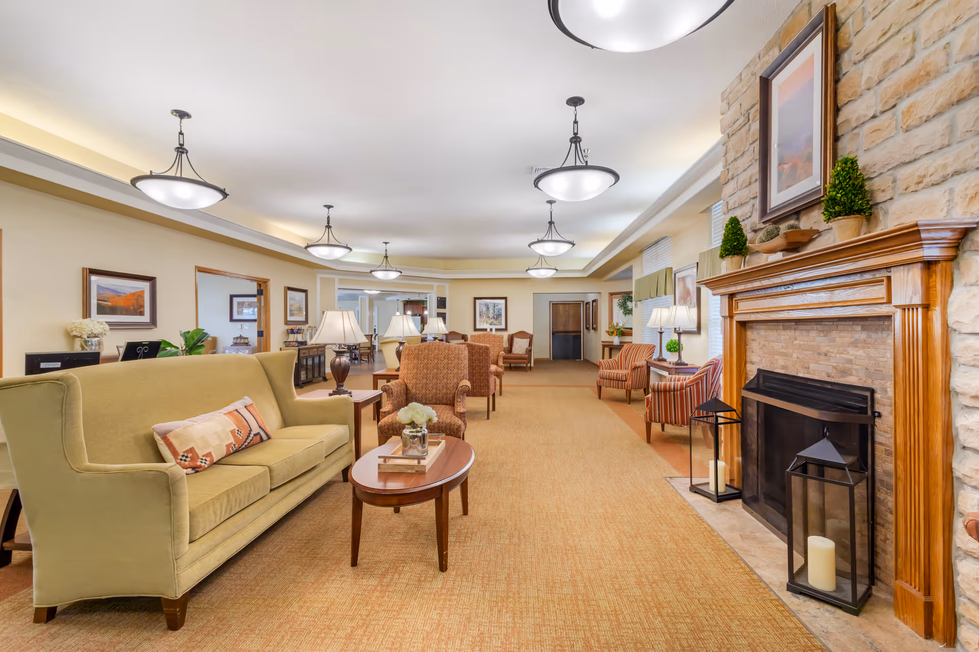 A spacious and well-lit living room area in a senior living facility featuring a green sofa with a decorative pillow, several upholstered armchairs, wooden side tables with lamps, and a stone fireplace with a wooden mantel decorated with small potted plants and a framed picture above it. The room has beige walls, carpeted flooring, and multiple ceiling lights.