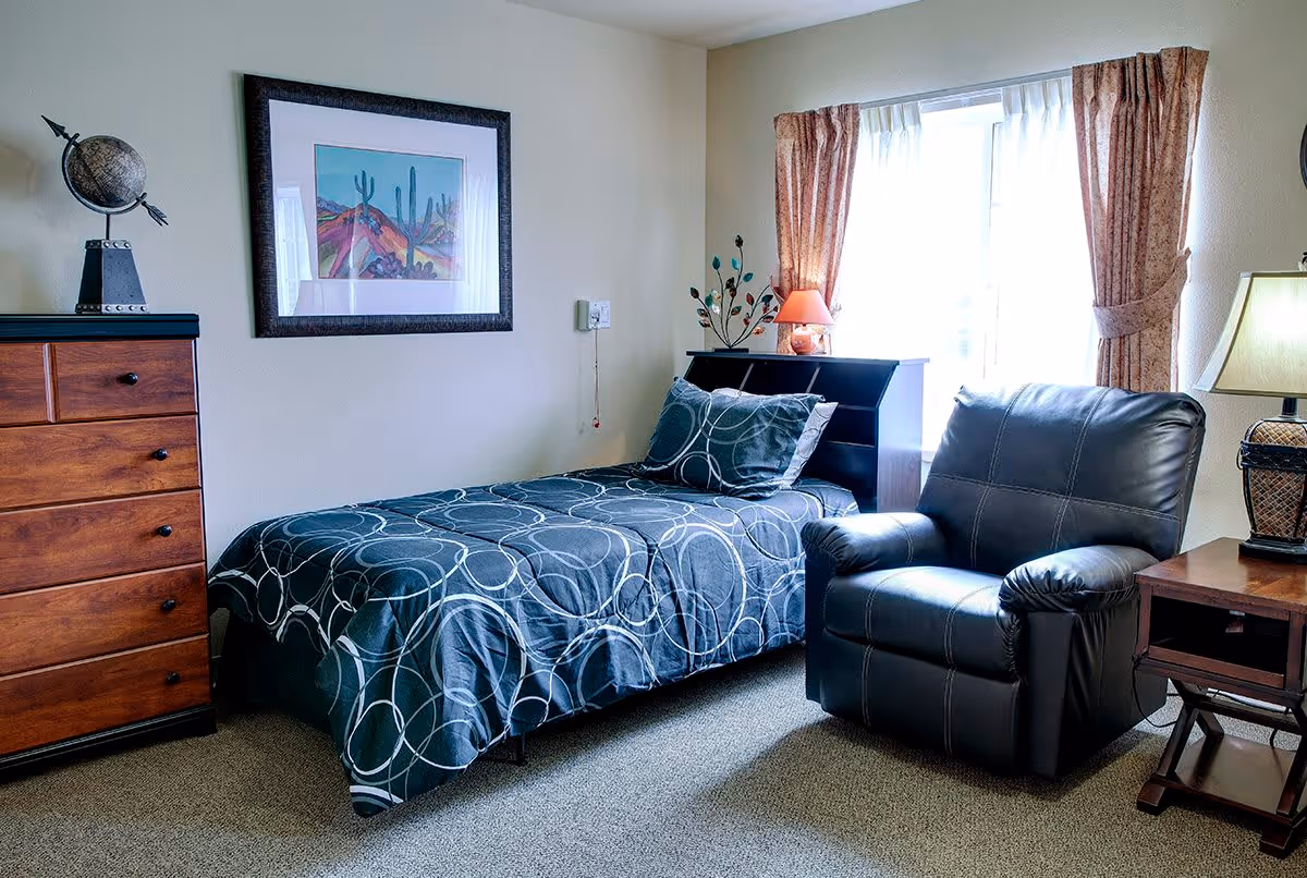 A cozy bedroom with a single bed covered in a dark blue bedspread with circular patterns. Next to the bed is a black leather armchair and a wooden side table with a lamp. A wooden dresser with a decorative globe on top is on the left side of the room. A framed painting of a desert landscape with cacti hangs on the wall above the bed. The room has a window with sheer white curtains and patterned drapes, allowing natural light to brighten the space.