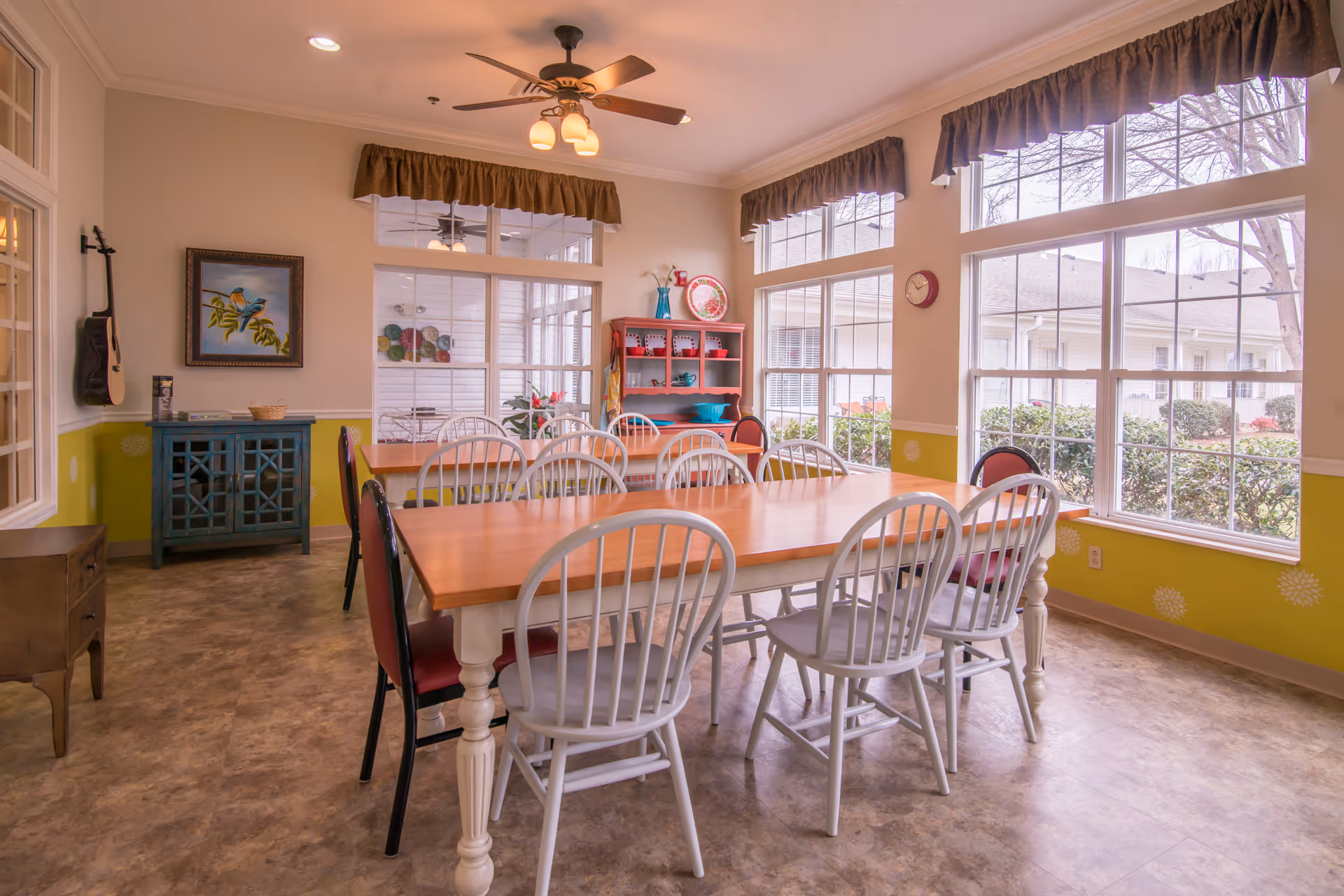A bright dining room with a long wooden table surrounded by white and red chairs. Large windows with brown valances let in natural light and offer a view of bushes and neighboring buildings outside. The room features a ceiling fan with lights, a blue cabinet with a painting of birds above it, and a red hutch displaying plates and decorative items.