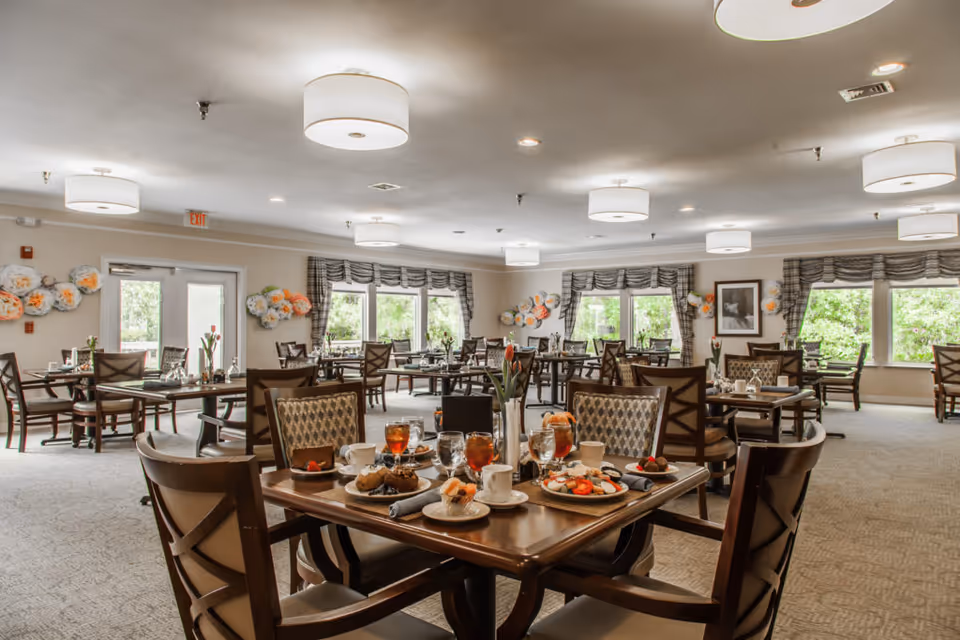 A spacious dining room with multiple wooden tables and chairs arranged neatly. Each table is set with plates of food, glasses of iced tea, coffee cups, and napkins. The room has large windows with patterned valances letting in natural light and decorated walls with large floral arrangements. Ceiling lights provide additional illumination.