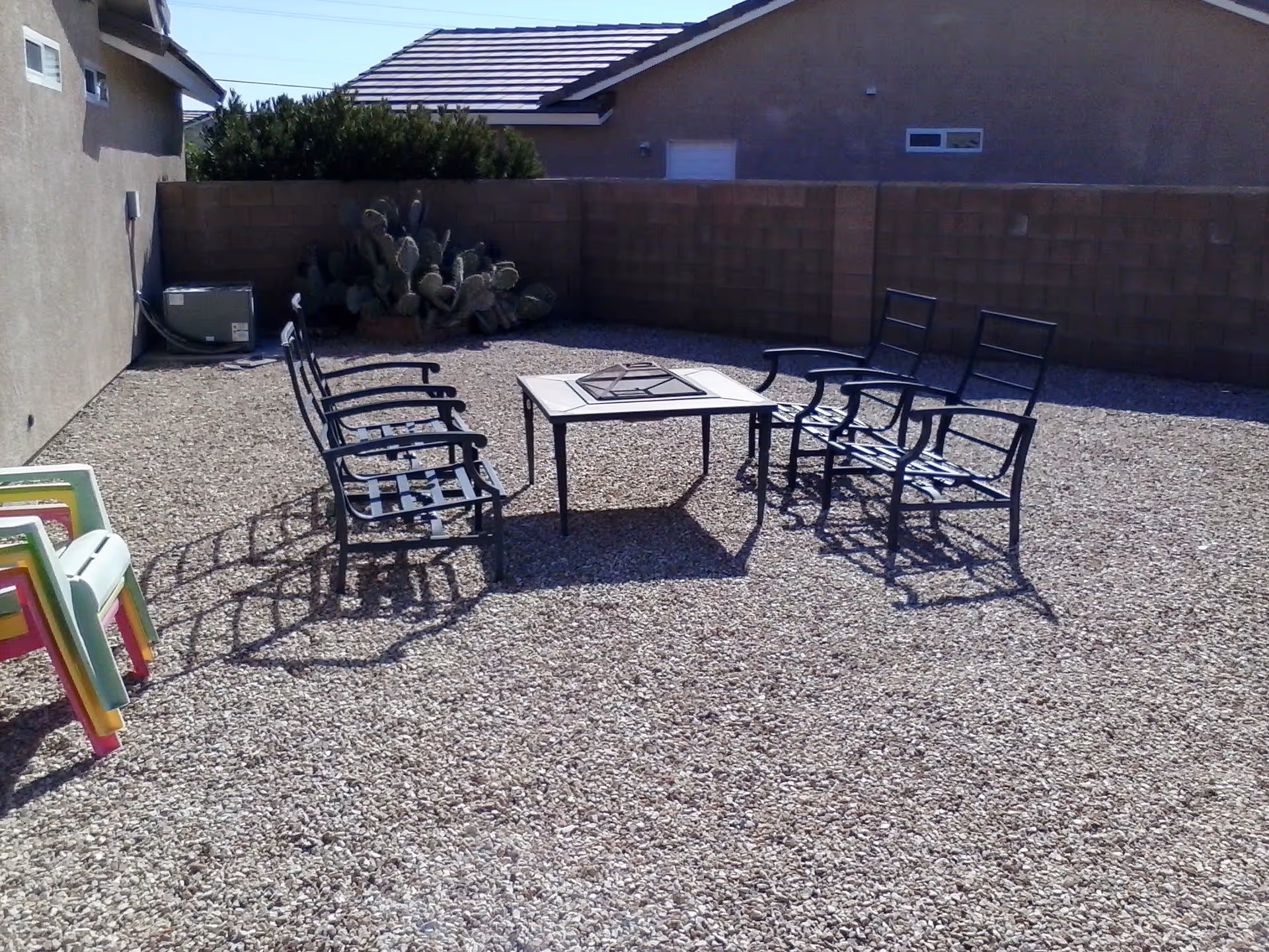 Outdoor seating area with two metal benches facing each other and a square fire pit table in the center, surrounded by gravel ground and enclosed by a brick wall. There are some colorful plastic chairs stacked on the left side and a cactus plant near the back wall.