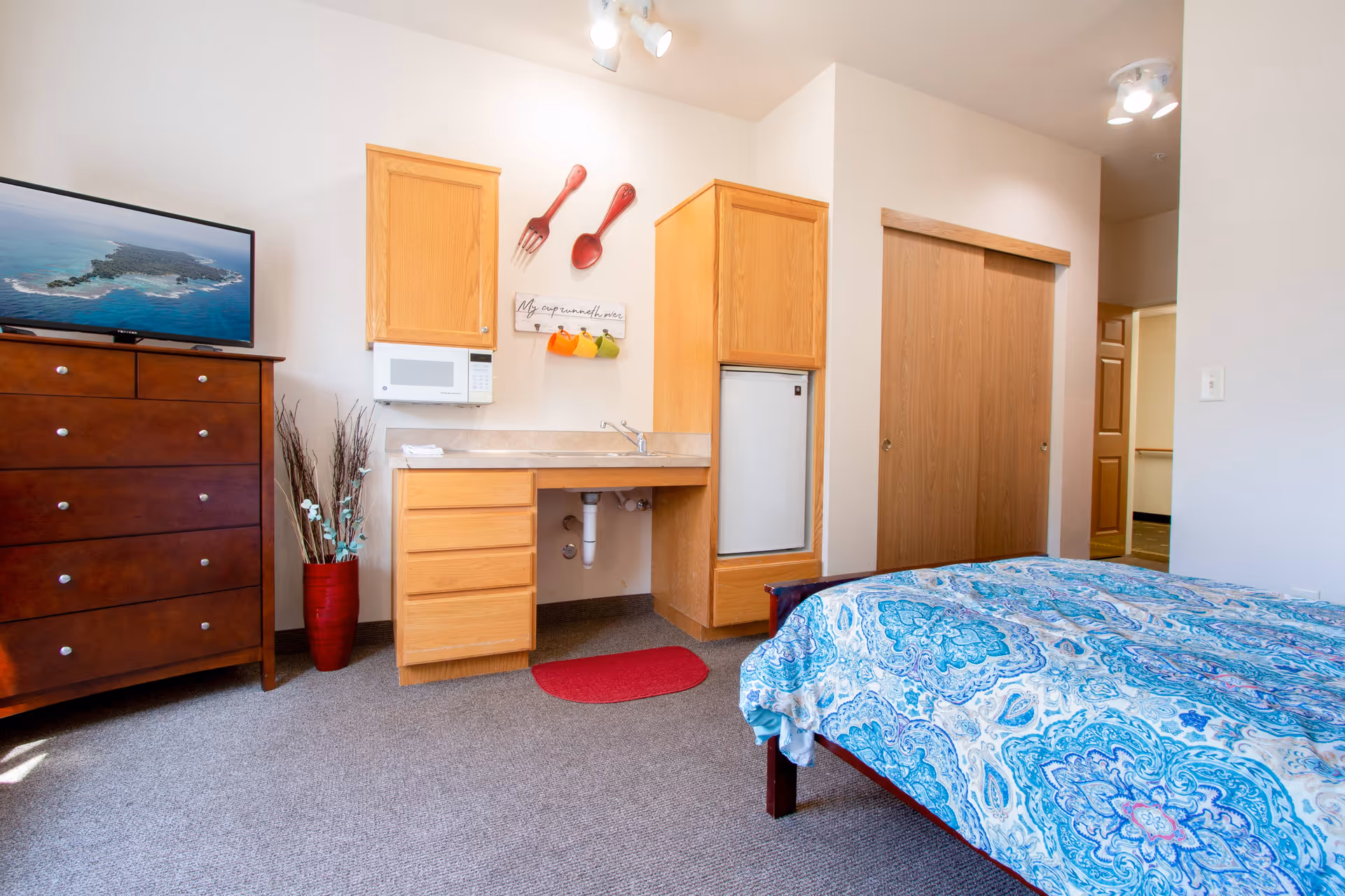 A senior living facility room featuring a bed with a blue patterned bedspread, a wooden dresser with a flat-screen TV on top, a small kitchenette area with wooden cabinets, a microwave, a sink, and a mini refrigerator. There is a red rug on the carpeted floor and decorative oversized fork and spoon wall hangings above the kitchenette. A closet with sliding wooden doors is visible in the background.