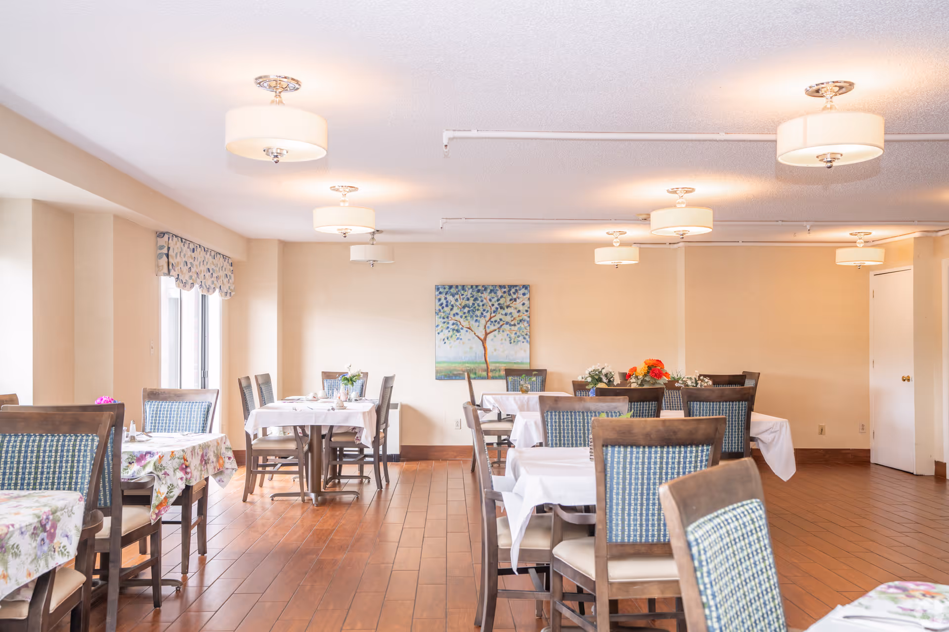 A bright dining room with several tables covered in white and floral tablecloths, surrounded by wooden chairs with blue patterned cushions. The room has wooden flooring, beige walls, ceiling lights, and a painting of a tree on the far wall. Windows on the left side allow natural light to enter.