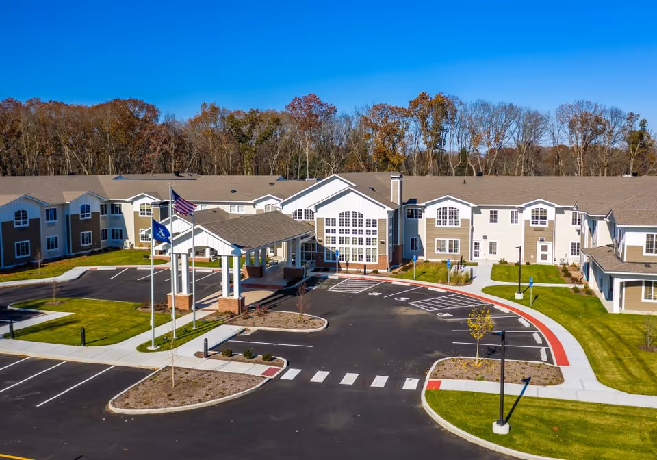 Front entrance of a two-story senior living facility with a porte-cochère, flagpoles, parking lot, and landscaped grounds under a clear blue sky.