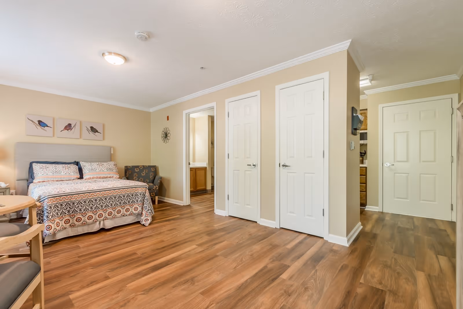 A spacious bedroom in a senior living facility with a bed featuring patterned bedding, a cushioned armchair with floral upholstery, and three bird-themed artworks on the wall above the bed. The room has light beige walls, wood flooring, and multiple white doors leading to other rooms, including a bathroom and a kitchen area.