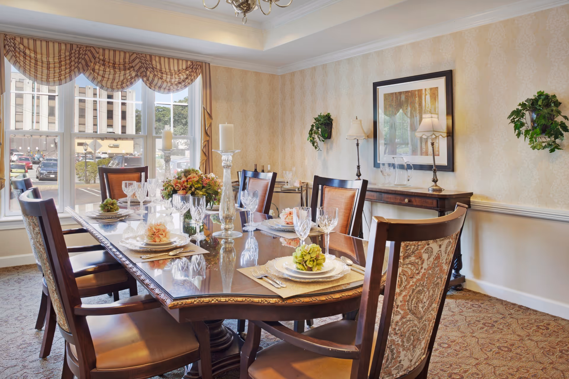 A formal dining room with a set wooden table and chairs, place settings and centerpieces, large windows, and a sideboard against the wallpapered wall.