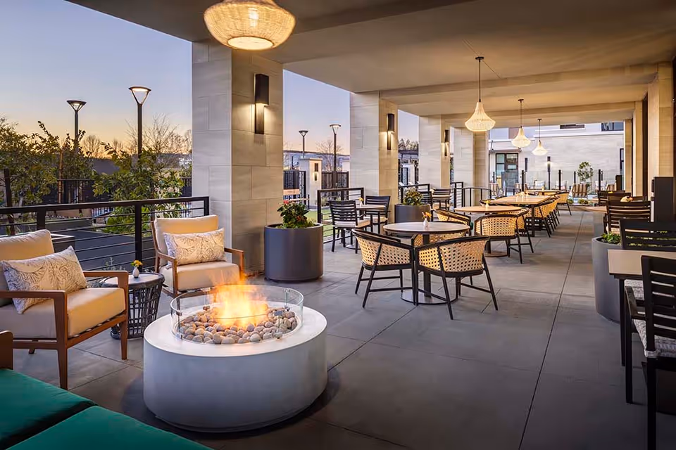 Outdoor covered patio area at Belmont Village San Ramon with multiple tables and chairs arranged for seating. There are cushioned armchairs around a circular fire pit with flames, large planters with greenery, and modern hanging light fixtures. The setting is during dusk with a clear sky and some outdoor lighting visible.