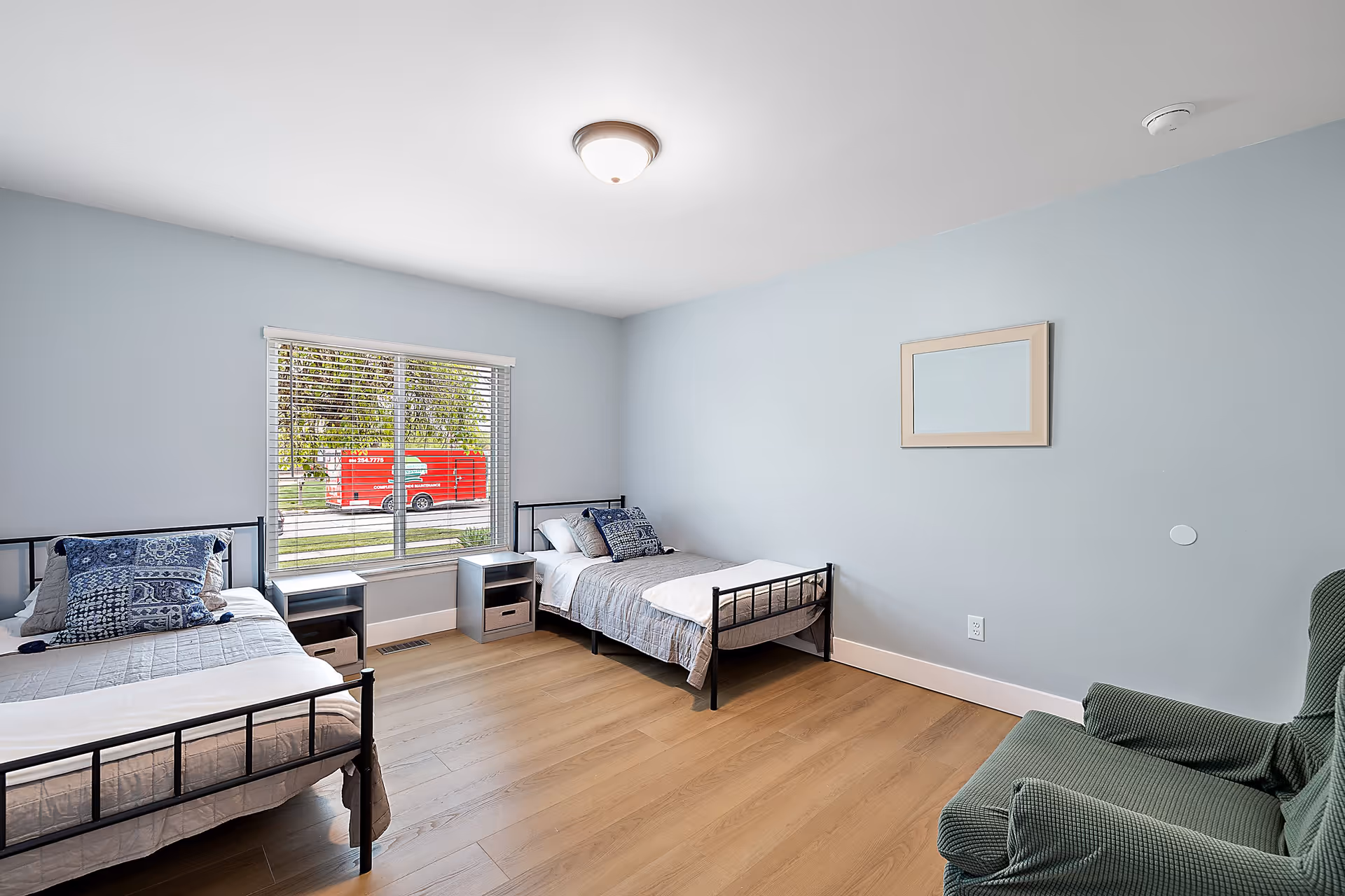 A simple bedroom with two single beds, each with a blue patterned pillow and gray bedding. Between the beds are two small nightstands with storage baskets. A large window with white blinds lets in natural light and shows a red vehicle outside. The walls are painted light blue, and there is a green armchair in the corner. A rectangular mirror hangs on the wall above an electrical outlet.