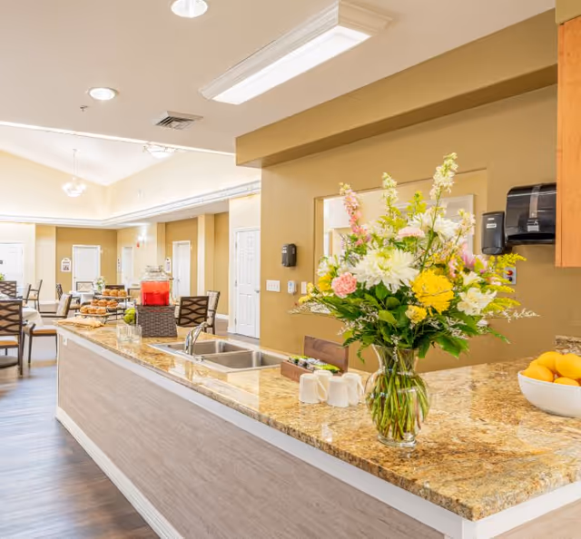 Bright communal dining and serving area with a granite countertop island, a vase of flowers, a sink, and tables and chairs in the background.