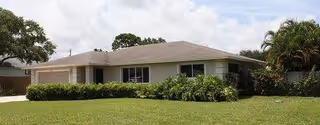Single-story residential building with a light-colored exterior, surrounded by green grass, bushes, and trees under a partly cloudy sky.
