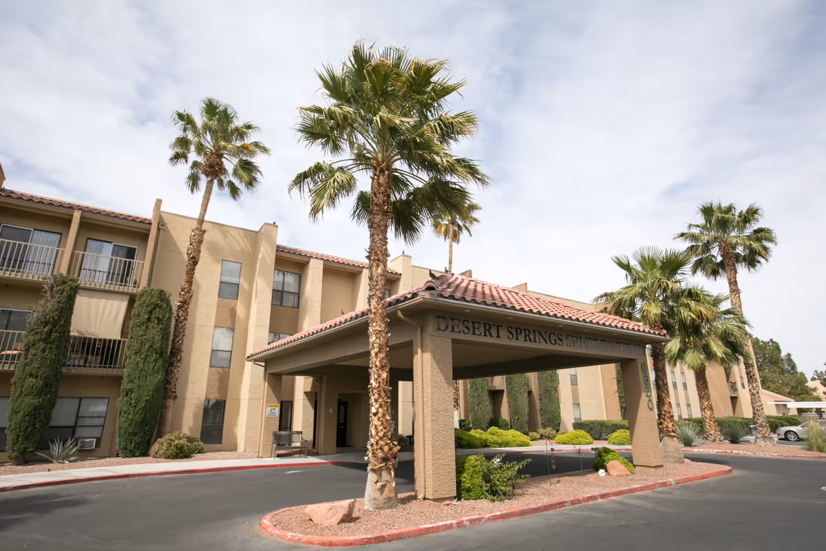 Exterior view of Desert Springs Senior Living facility with a covered entrance, palm trees, and a three-story beige building under a partly cloudy sky.
