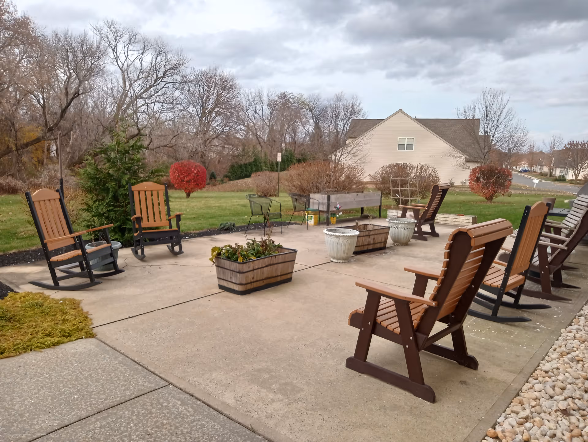 Outdoor patio area with several wooden rocking chairs and planters on a concrete surface, surrounded by grass and trees with some bushes showing autumn colors. A house and a road are visible in the background under a cloudy sky.