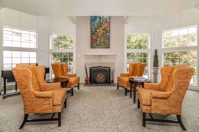 Sunlit sitting room with six orange wingback chairs arranged around a central fireplace beneath a colorful painting.