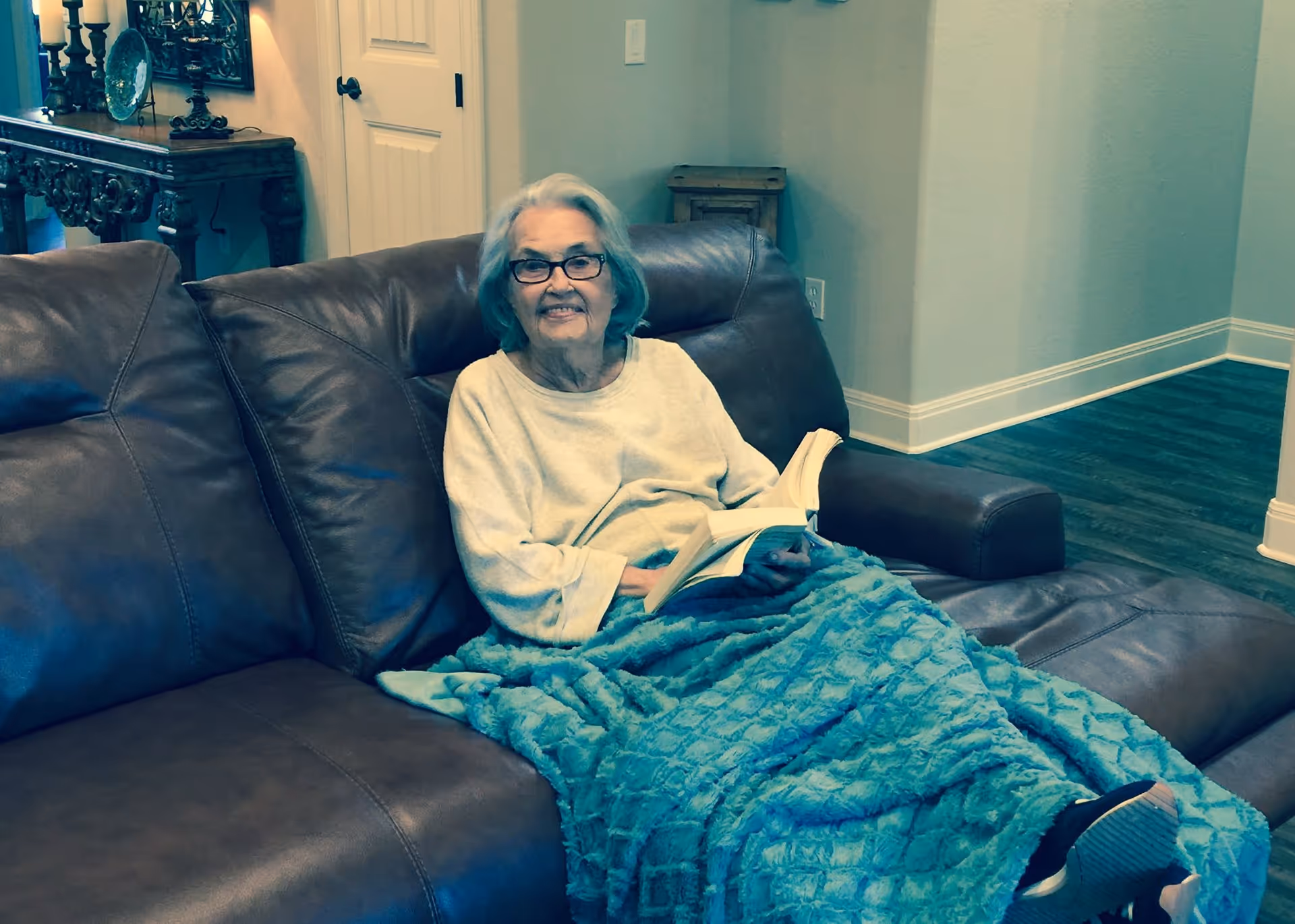 An elderly woman with glasses sitting on a brown leather couch, covered with a blue textured blanket, holding an open book and smiling. The room has light-colored walls, wooden flooring, and a decorative wooden table with a mirror and ornaments in the background.