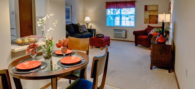 A cozy living and dining area in a senior living facility with a round glass dining table set for four with red plates and napkins, a small vase with flowers, a dark sofa with cushions, a red armchair, a window with red curtains, and warm lighting from table lamps.