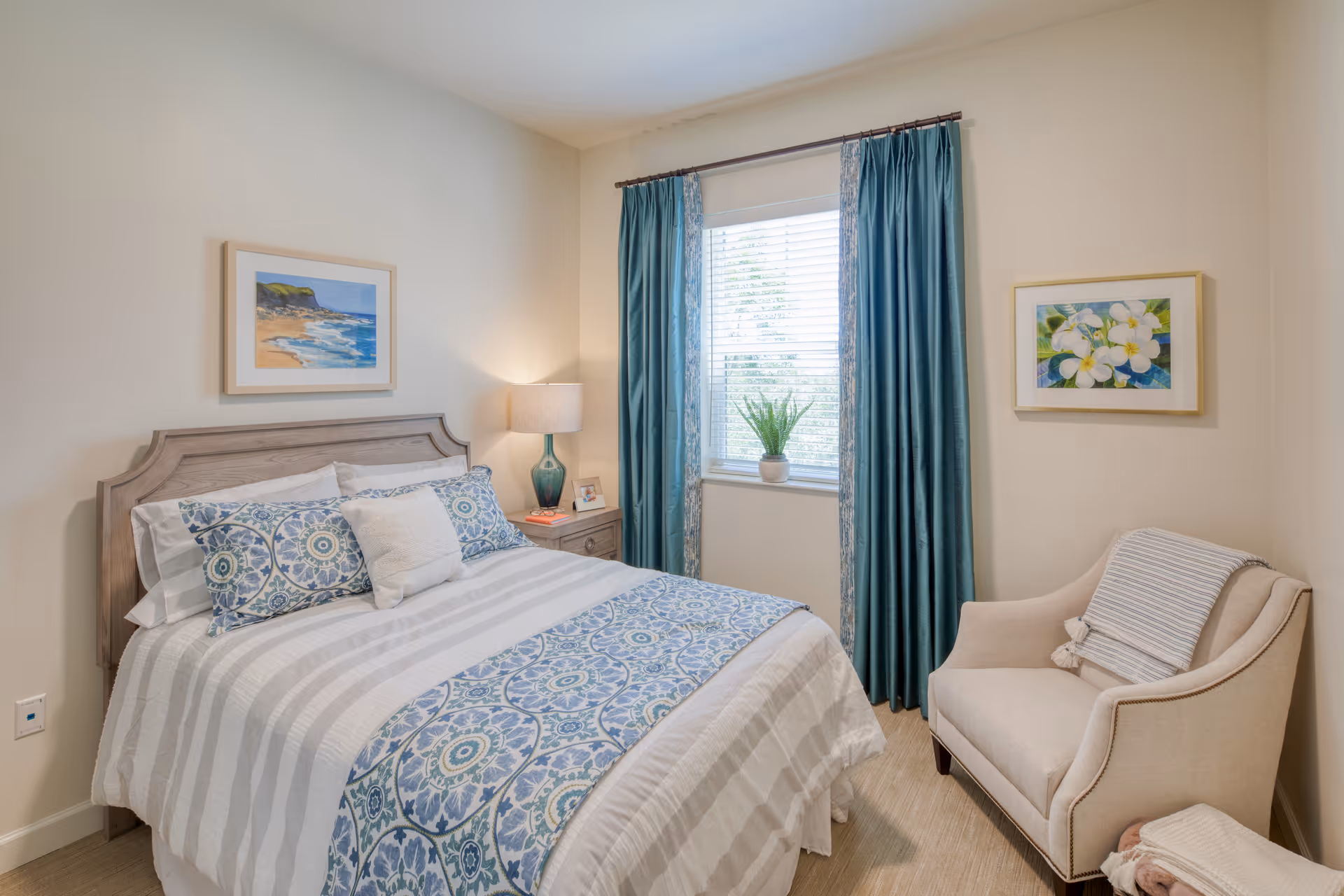 A cozy bedroom with a neatly made bed featuring patterned blue and white bedding. There is a wooden headboard, a nightstand with a lamp and a small framed photo, a window with blue curtains and a potted plant on the windowsill. A beige armchair with a striped throw blanket is positioned in the corner, and two framed pictures hang on the walls.